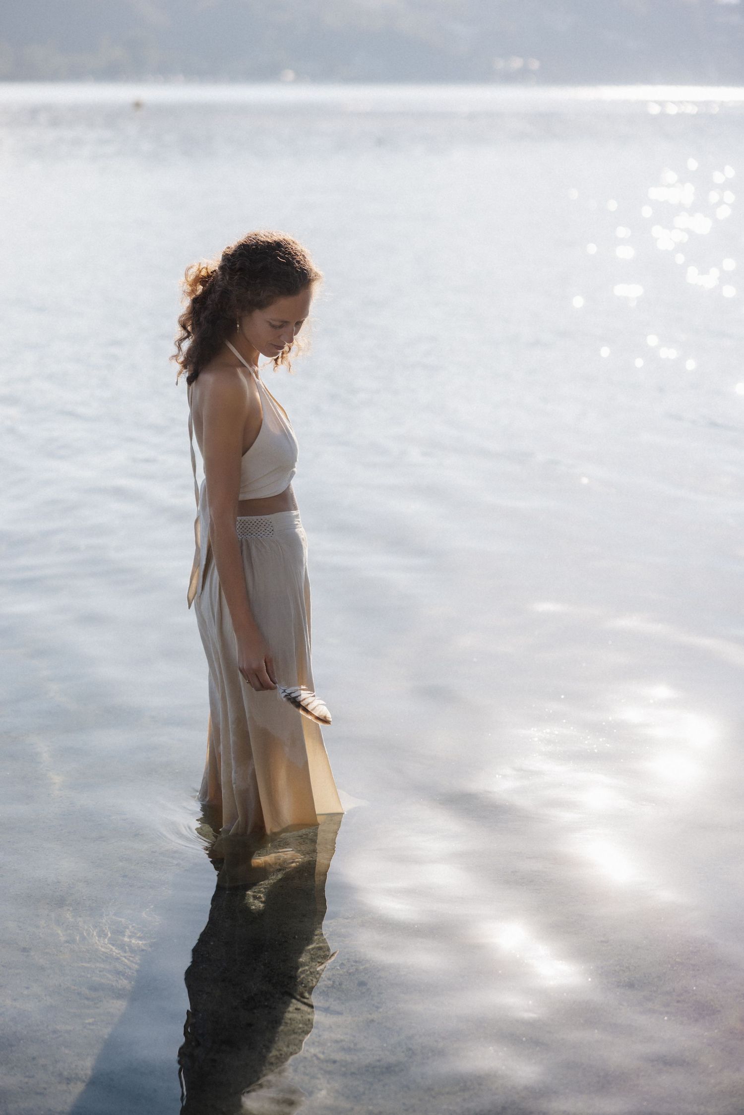 Curly-haired woman in cream two-piece outfit stands in shallow lake water holding a feather in sunlight.