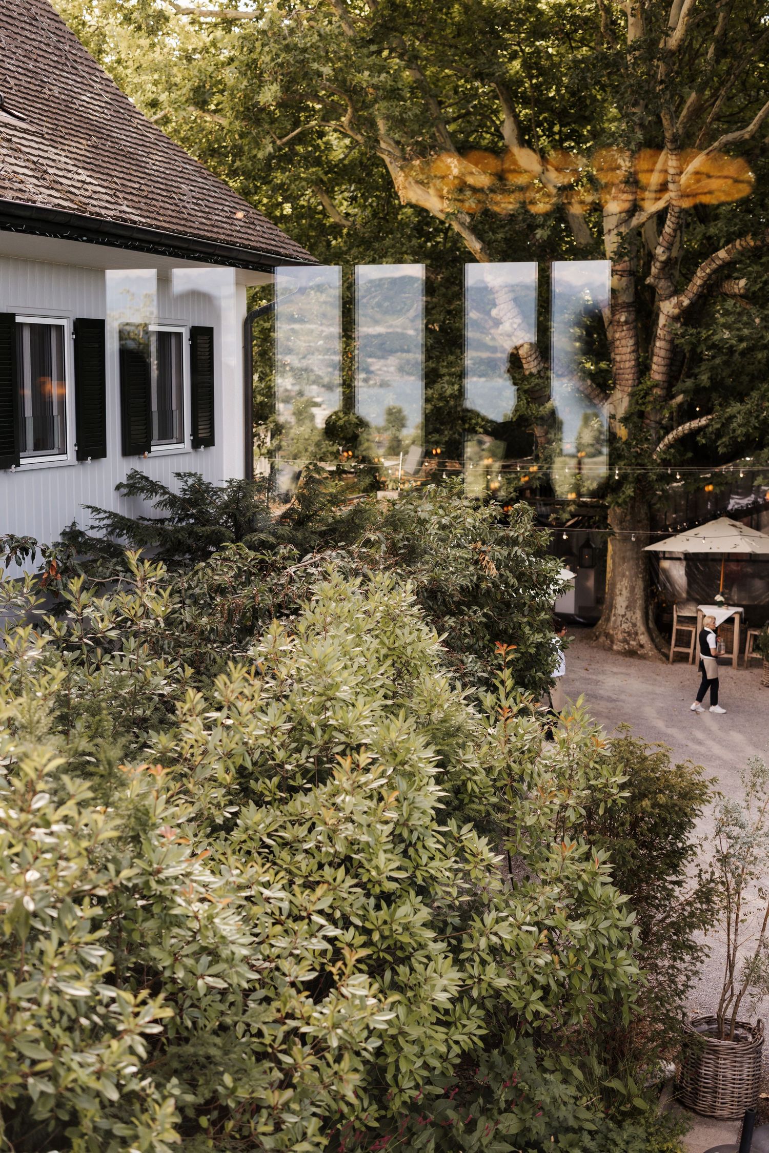 Aerial view of lush garden beside white country house with mountain landscape reflection.