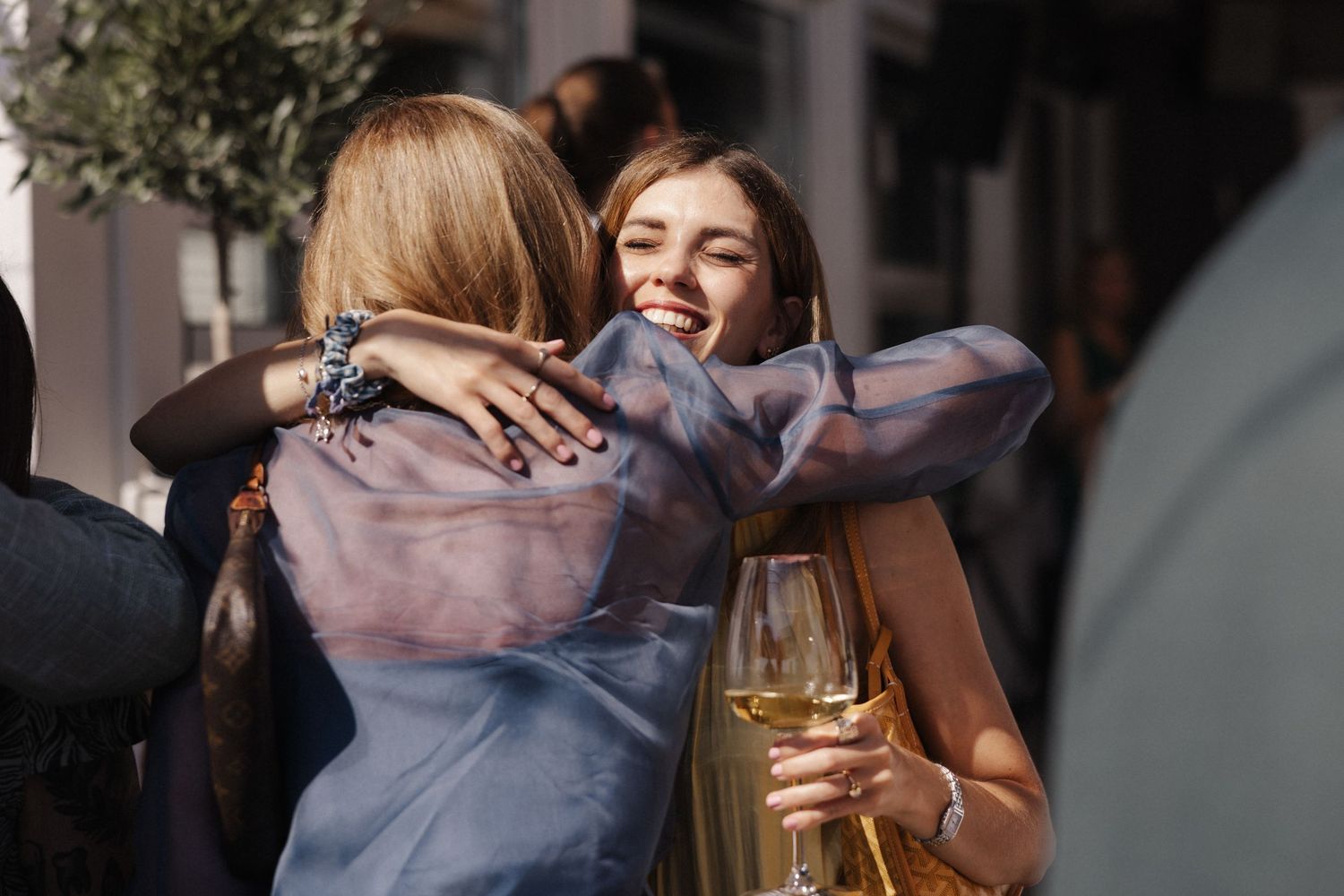 Two women sharing a warm embrace at an outdoor event, one holding a glass of white wine.