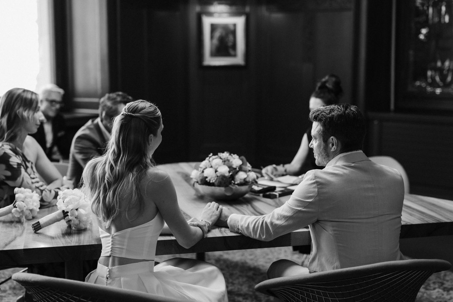 Black and white photo of bride and groom holding hands at ceremony table with guests and floral centerpiece nearby.