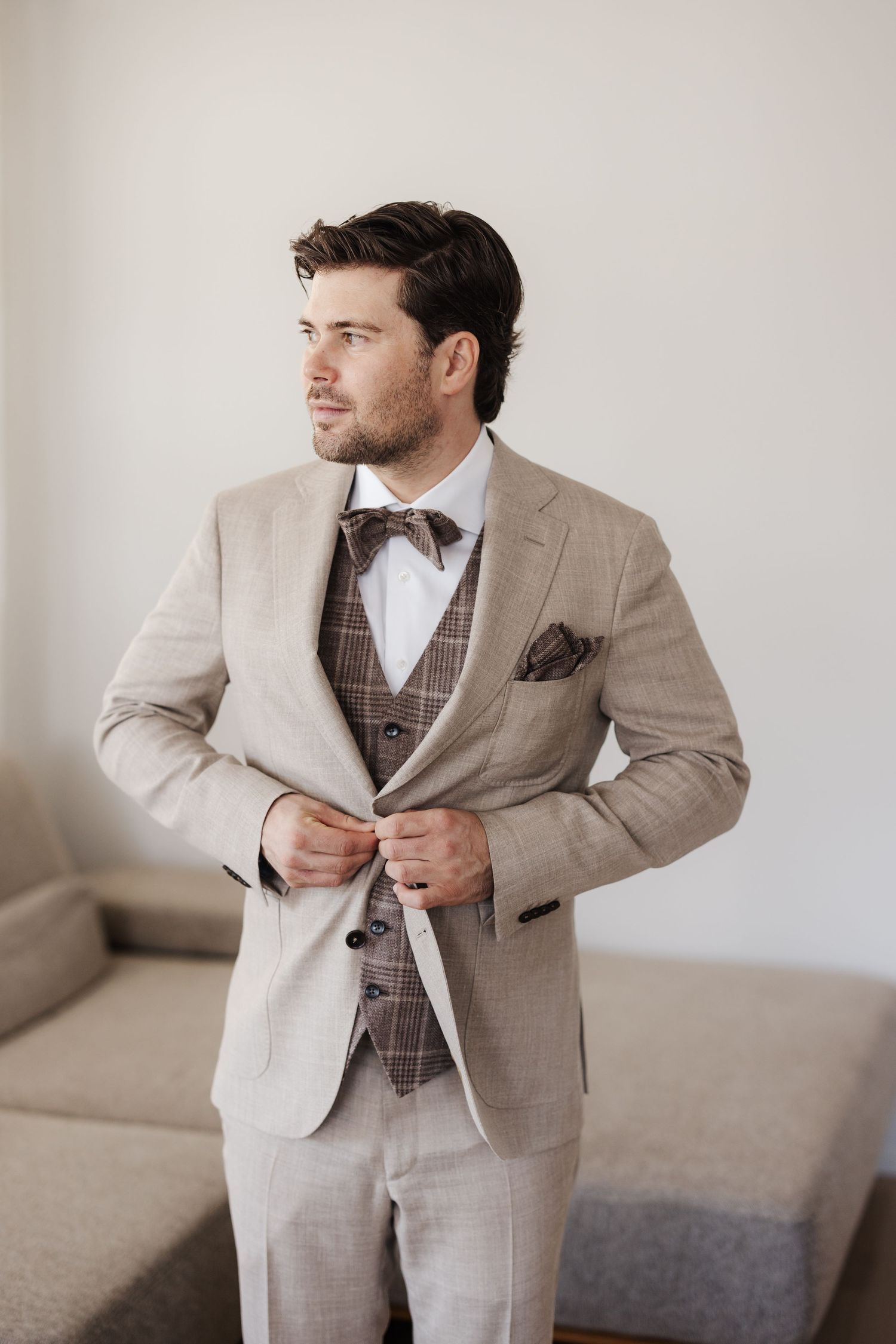 Handsome groom in a beige suit with brown plaid vest and bow tie buttoning his jacket indoors.
