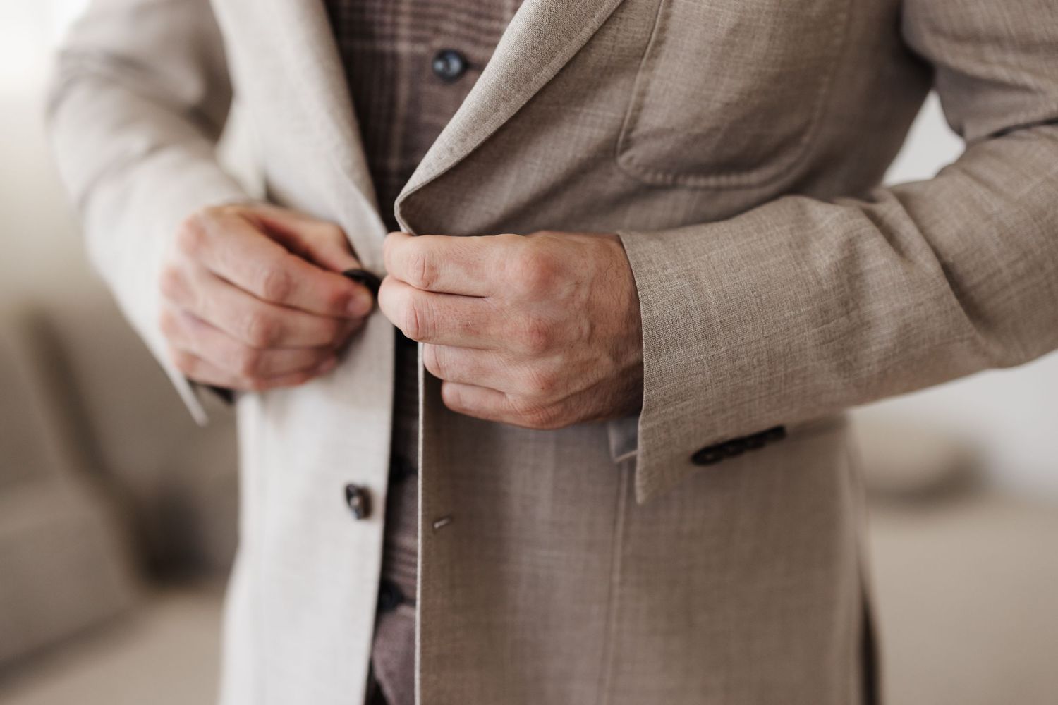 Close-up of a groom buttoning a light beige suit jacket over a brown checkered vest.