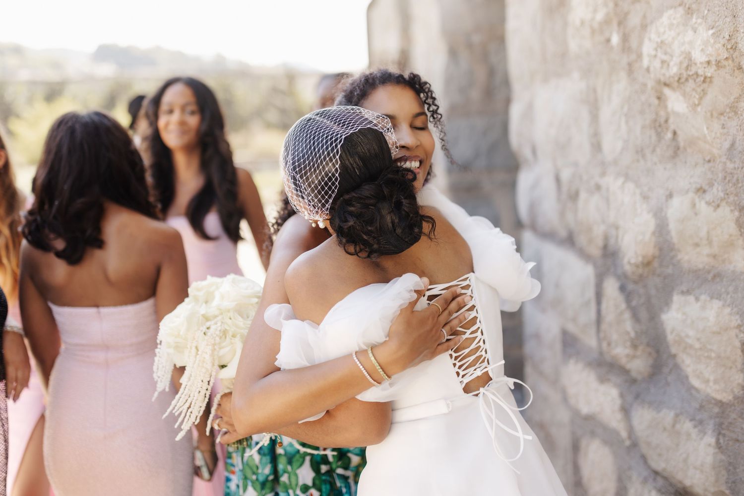 Bride in white off-shoulder gown with birdcage veil hugs guest while bridesmaids in pink stand nearby.