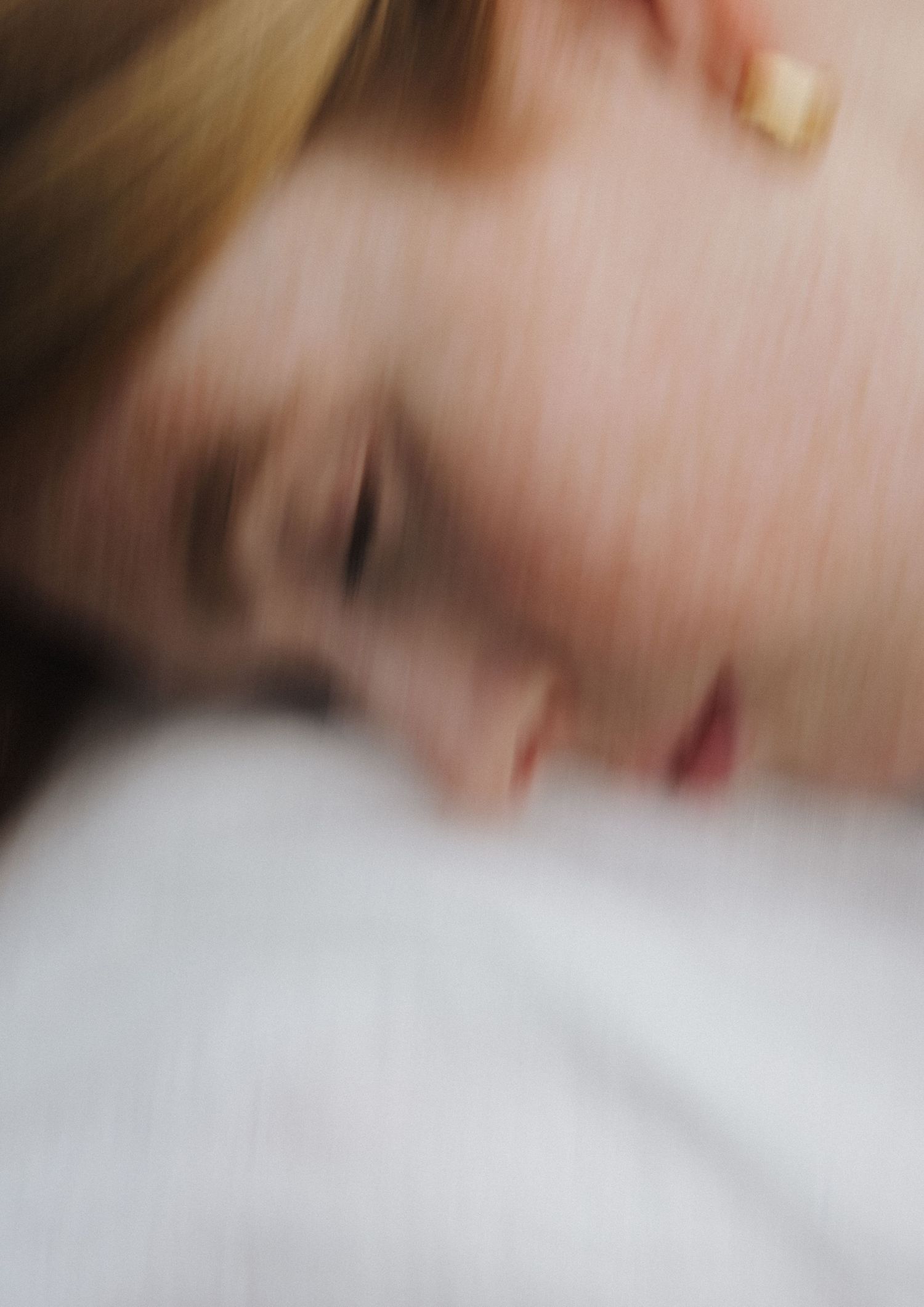 Soft blurred close-up of a sleeping woman with blonde hair and gold earring on white.