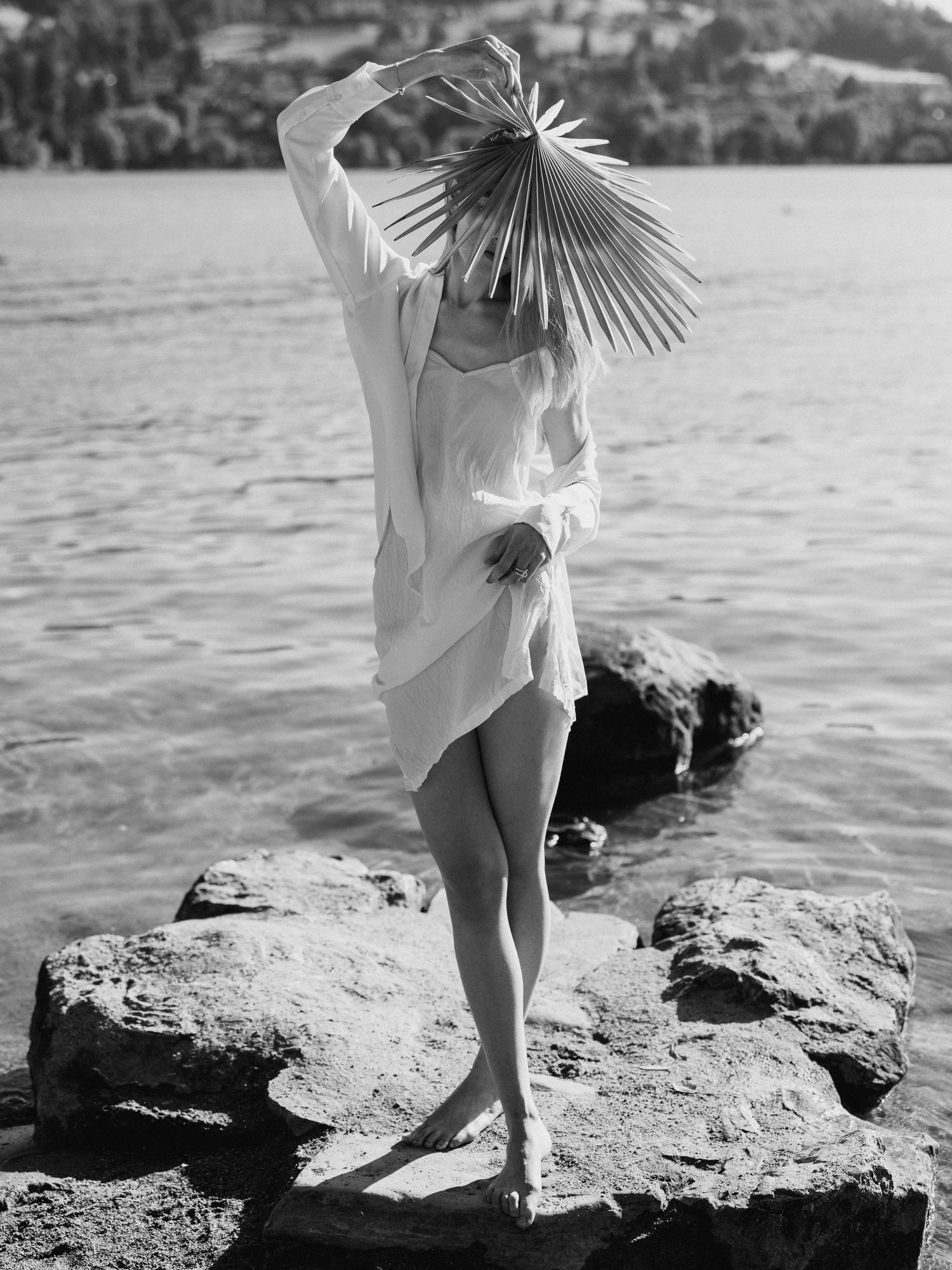 Black and white photo of woman holding fan over face on rocky lakeside shore.