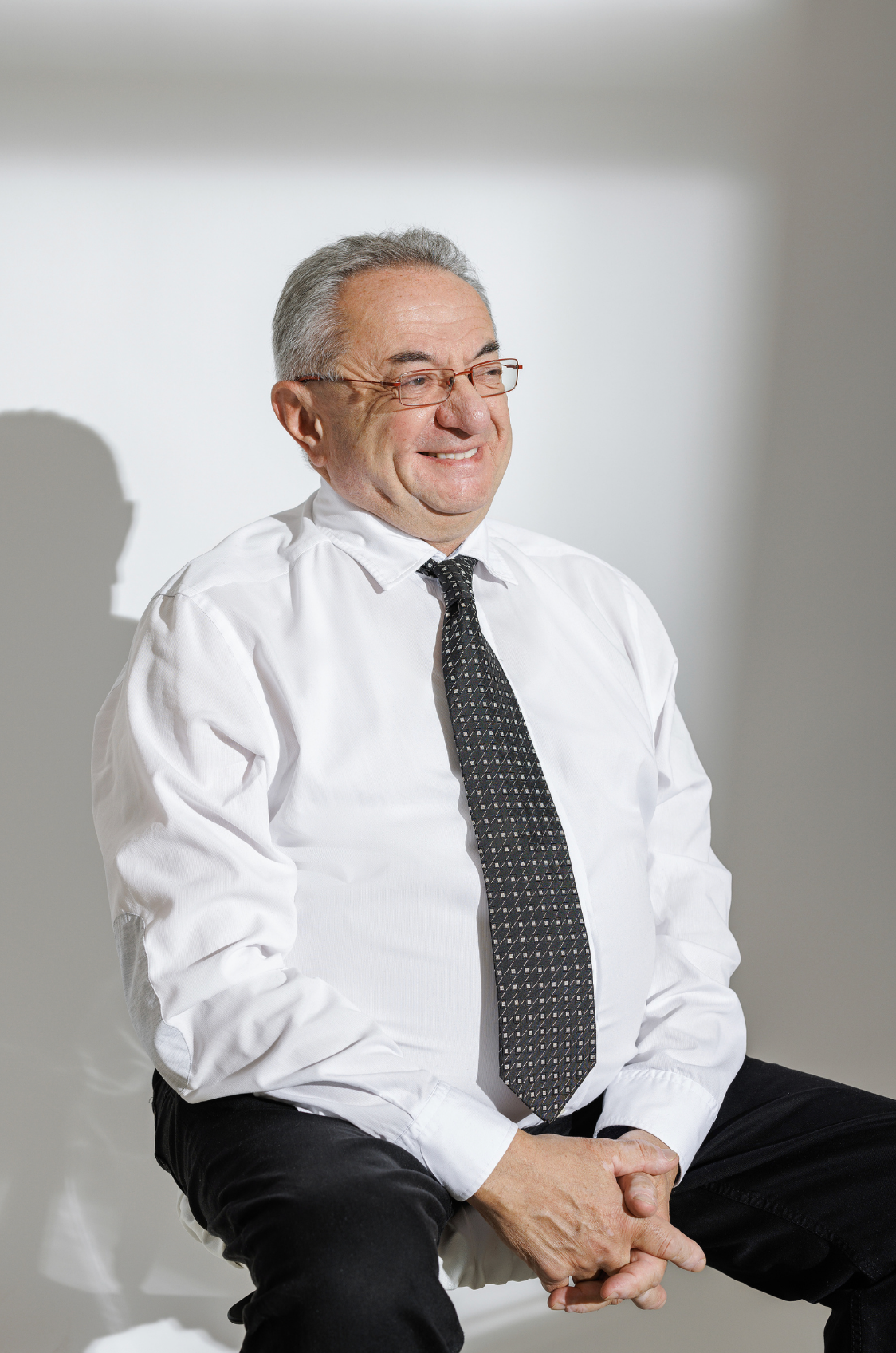 An older man in a white dress shirt and gray tie sits smiling with hands clasped.