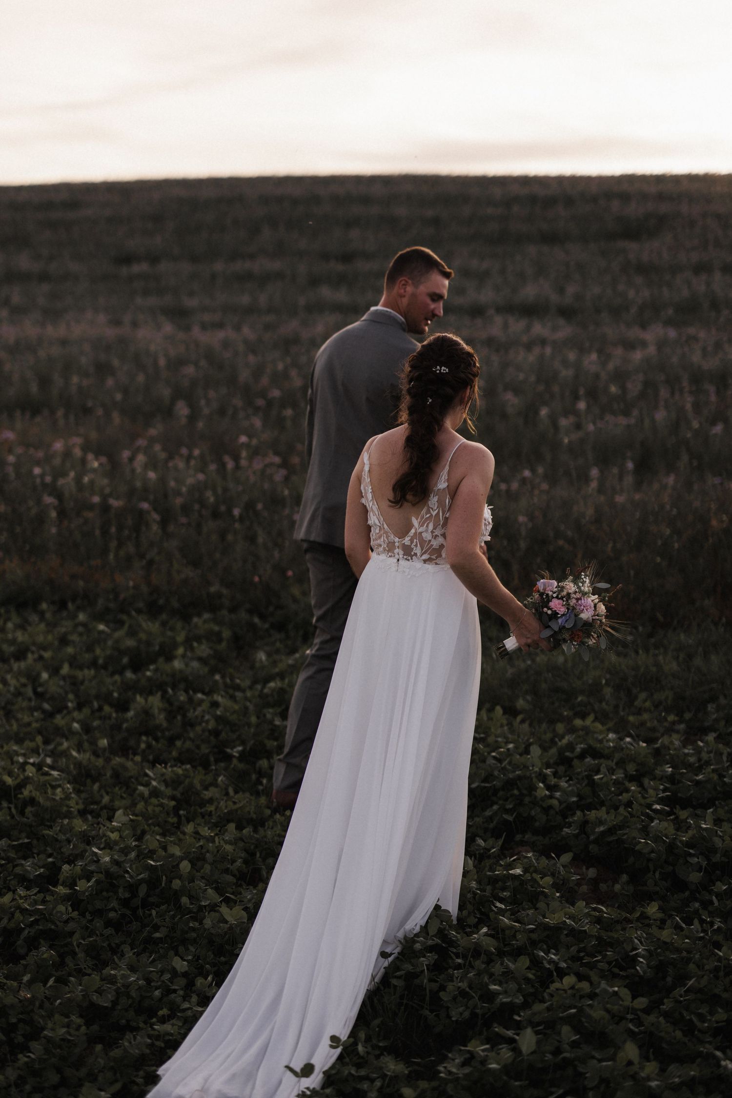 Bride in white lace gown holds bouquet while groom in gray suit looks at her in a field at sunset.