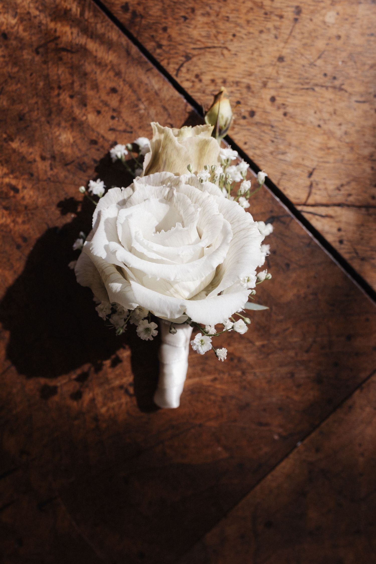 Elegant white rose boutonniere with baby's breath on rustic wooden parquet floor in natural light.