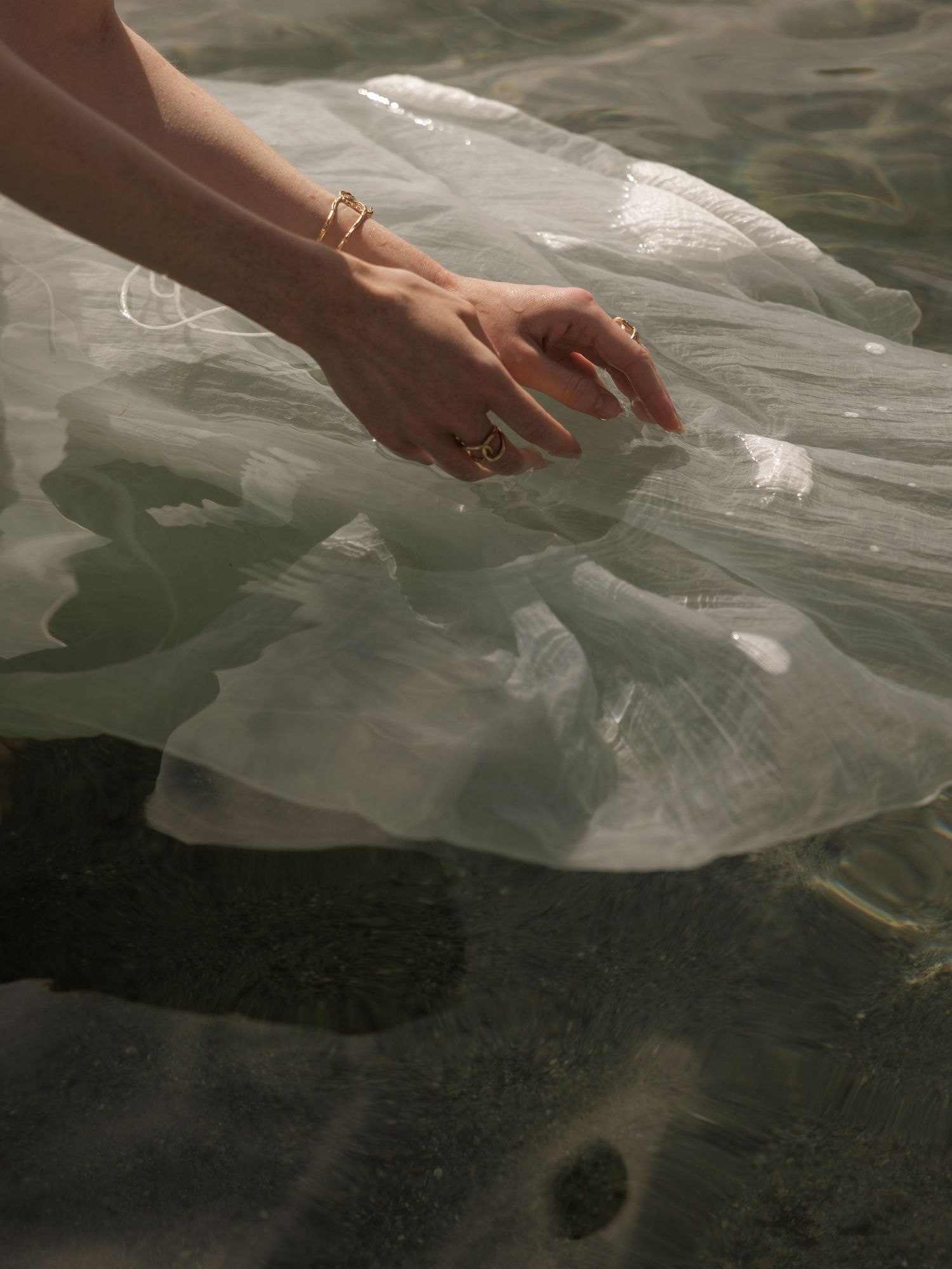Hand adorned with gold rings touching sheer white fabric floating on clear shallow water.