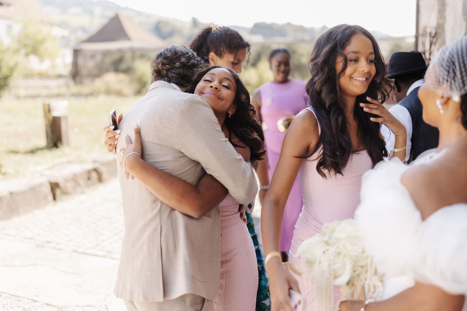 Wedding guests embrace and congratulate couple outdoors, bridesmaids in pink dresses holding white bouquets in sunlight.