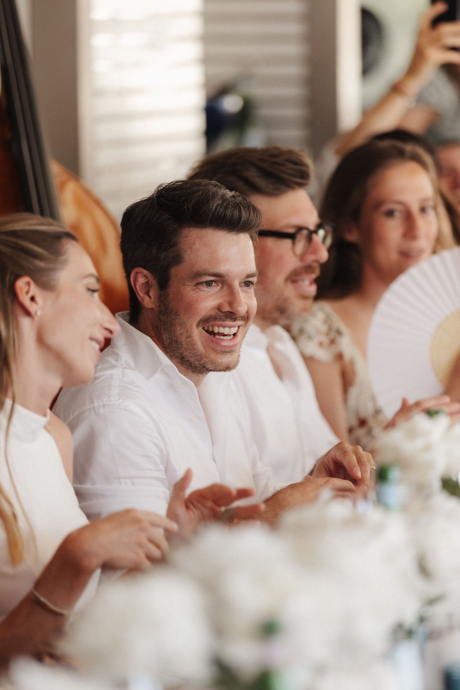 Smiling wedding guest in white shirt laughs with fellow guests holding decorative hand fans at reception.