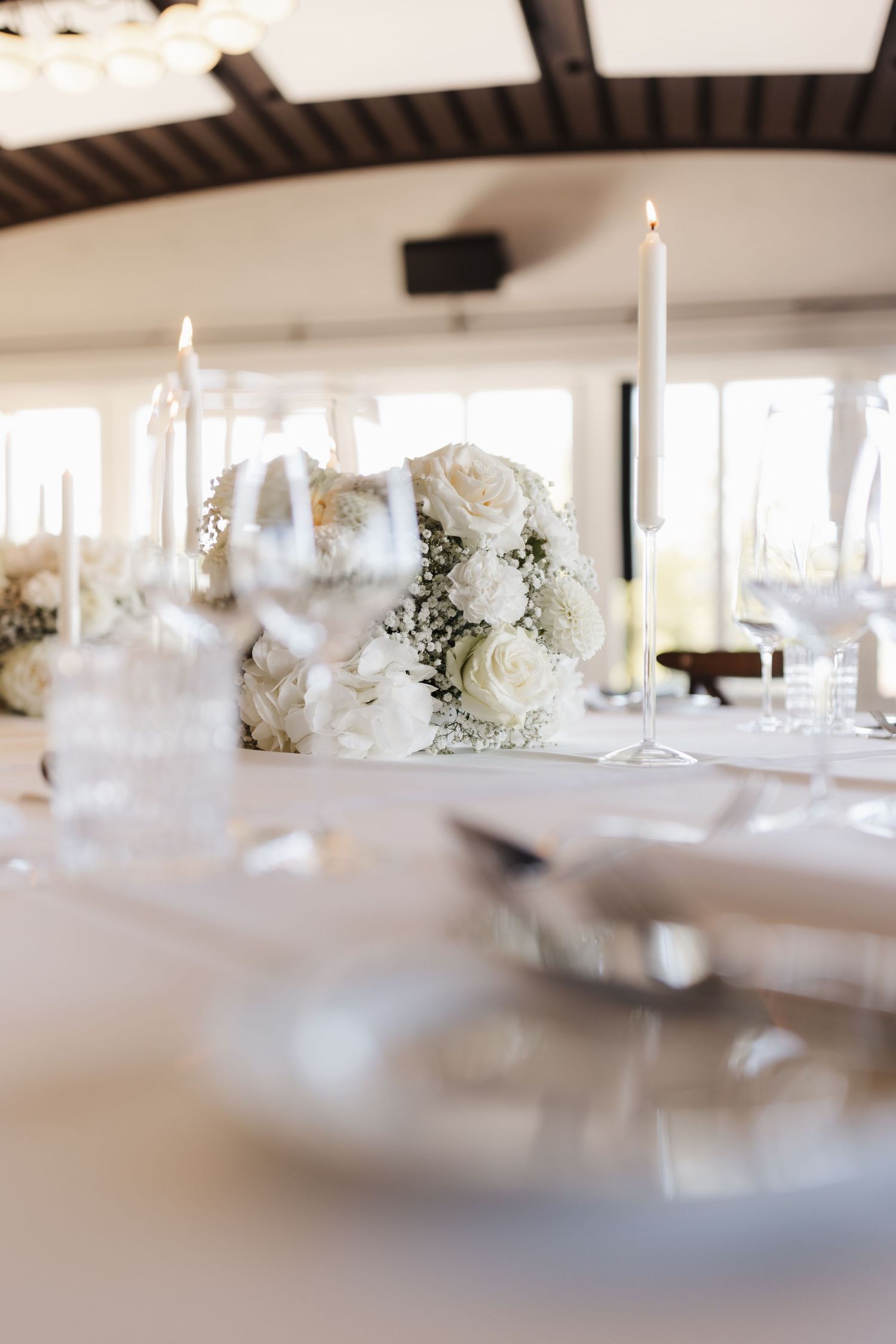 Elegant wedding reception table with white rose centerpiece, lit taper candles, crystal glassware and white linens.