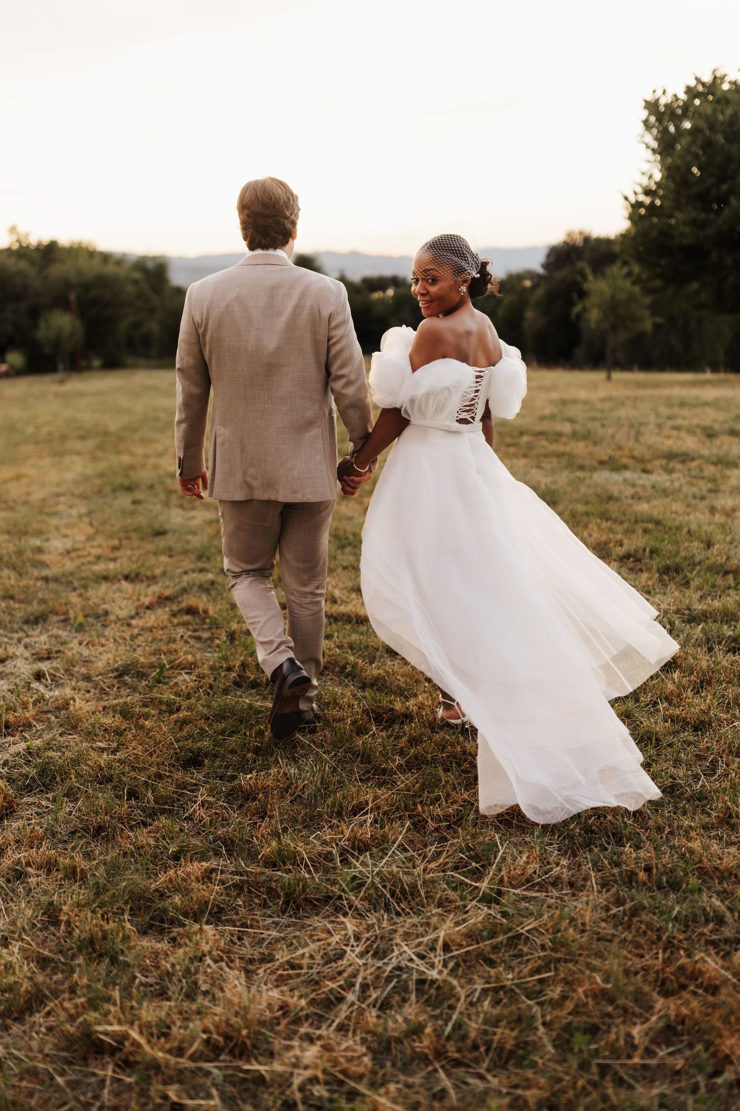 Bride in white puff-sleeve gown glances back while walking with groom through rustic meadow.
