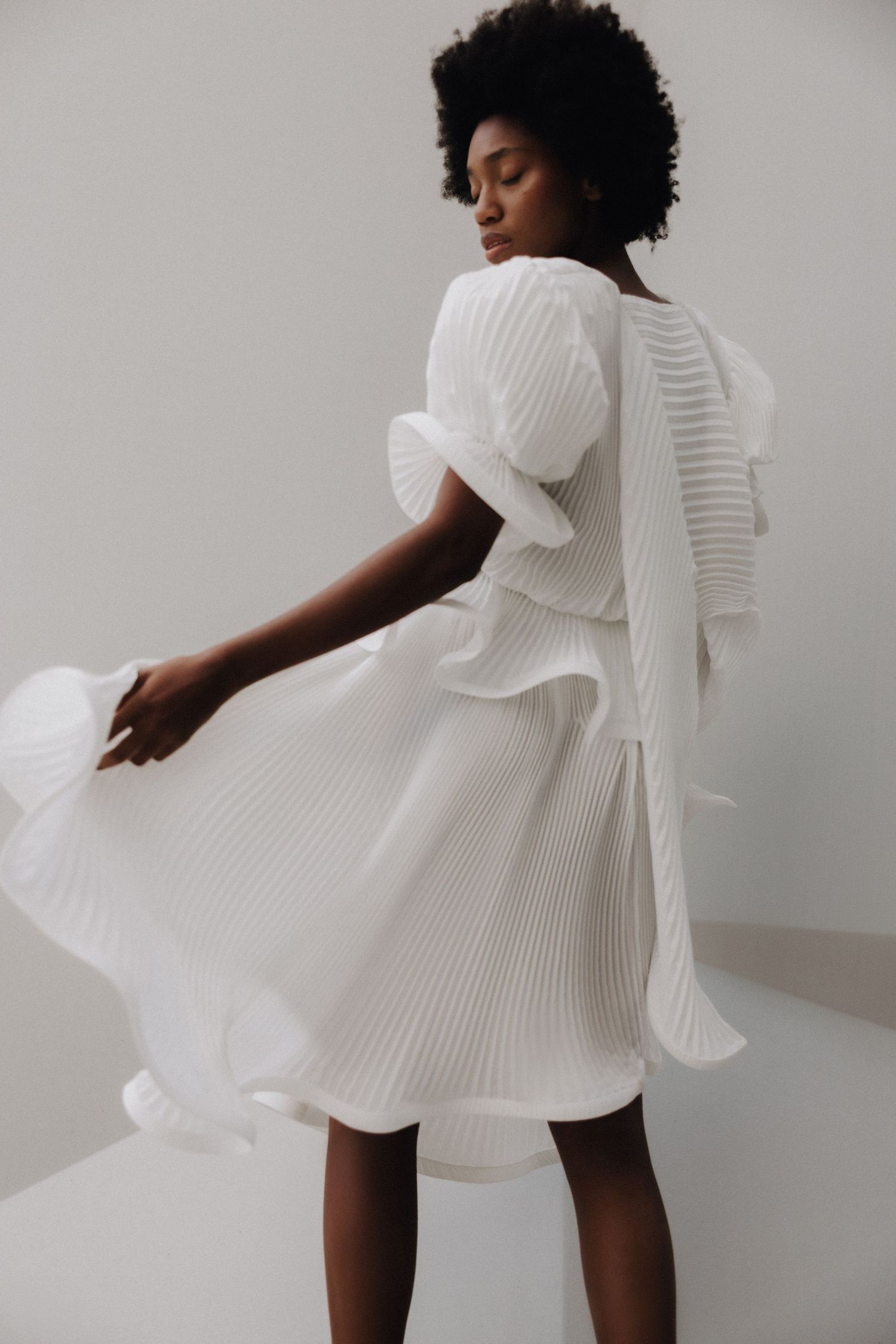 A Black woman twirls in a white pleated midi dress with ruffled back detail in a bright studio setting.