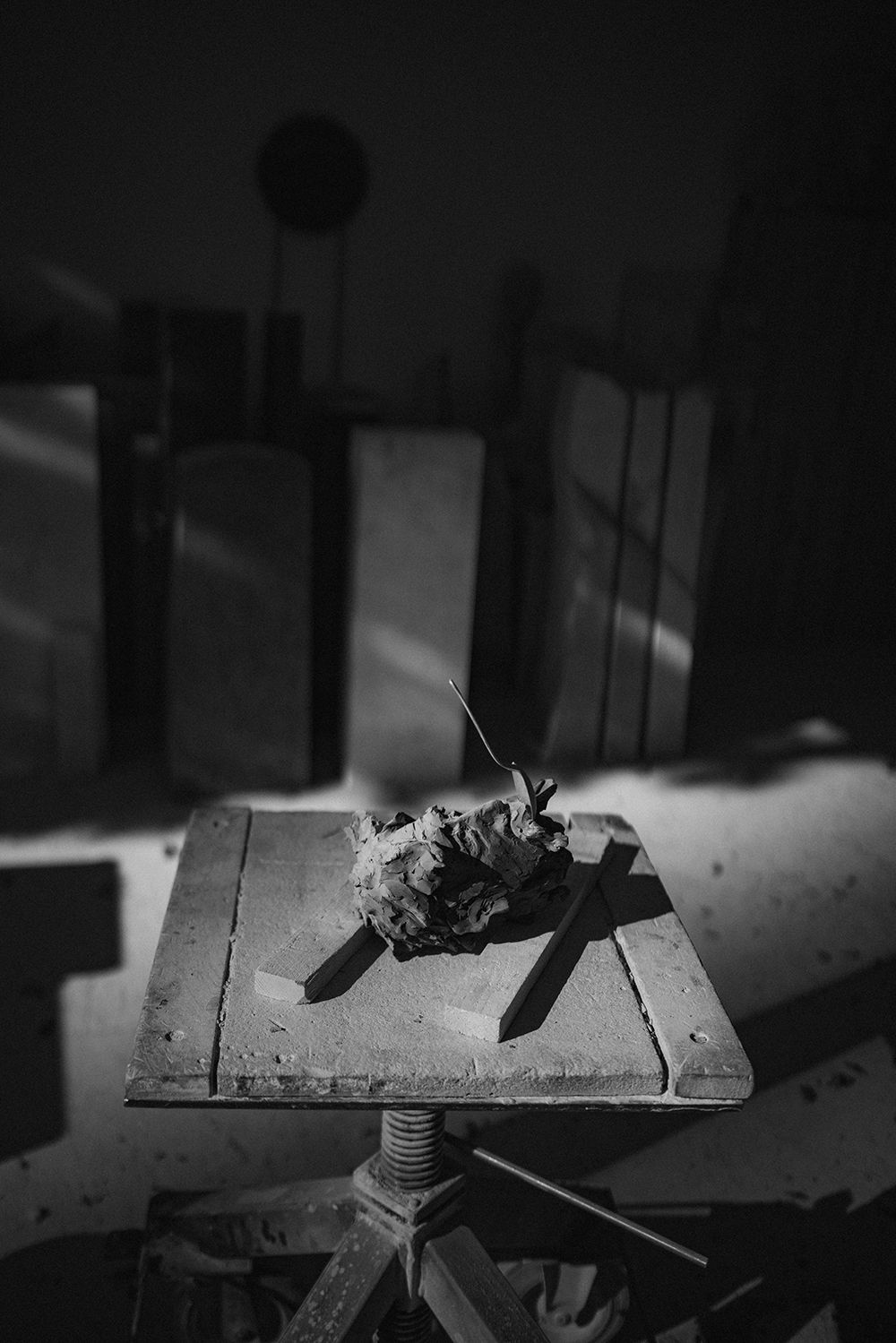 Black and white photo of a rough clay sculpture on a wooden adjustable stand in a dark studio.