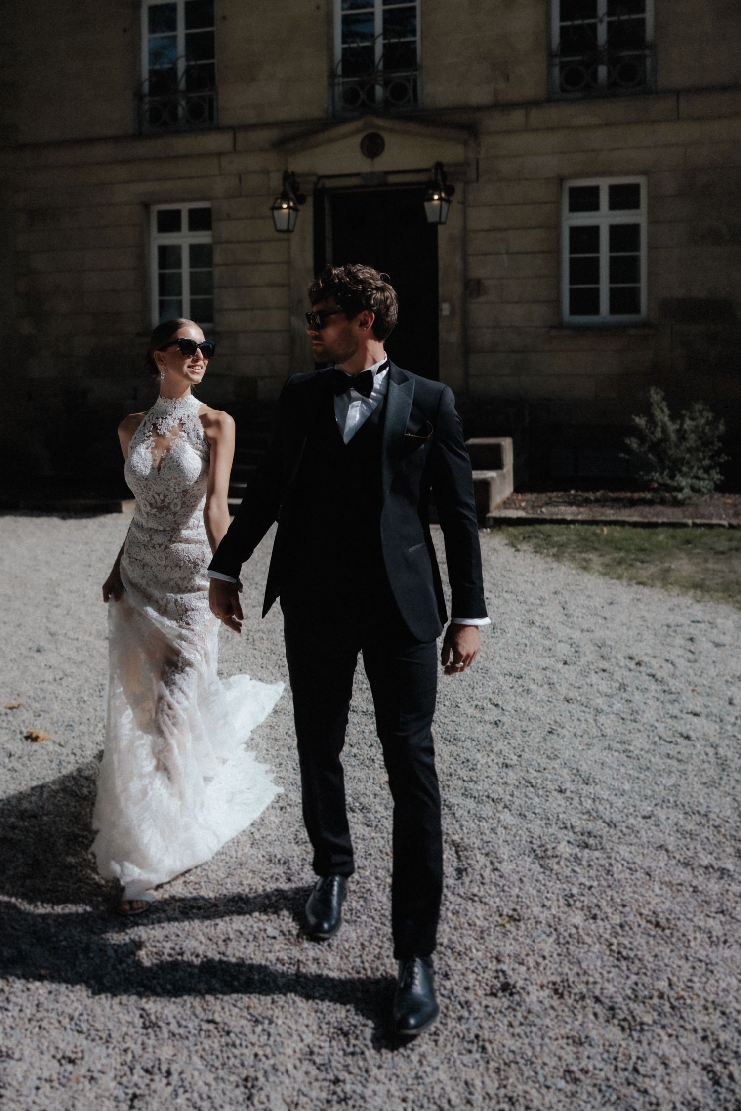 Stylish bride and groom in sunglasses walk hand in hand on gravel path before a historic manor.