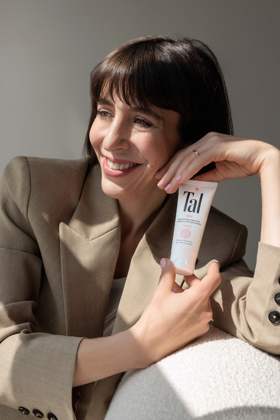 Smiling brunette woman in beige blazer holding Tal skincare product tube.