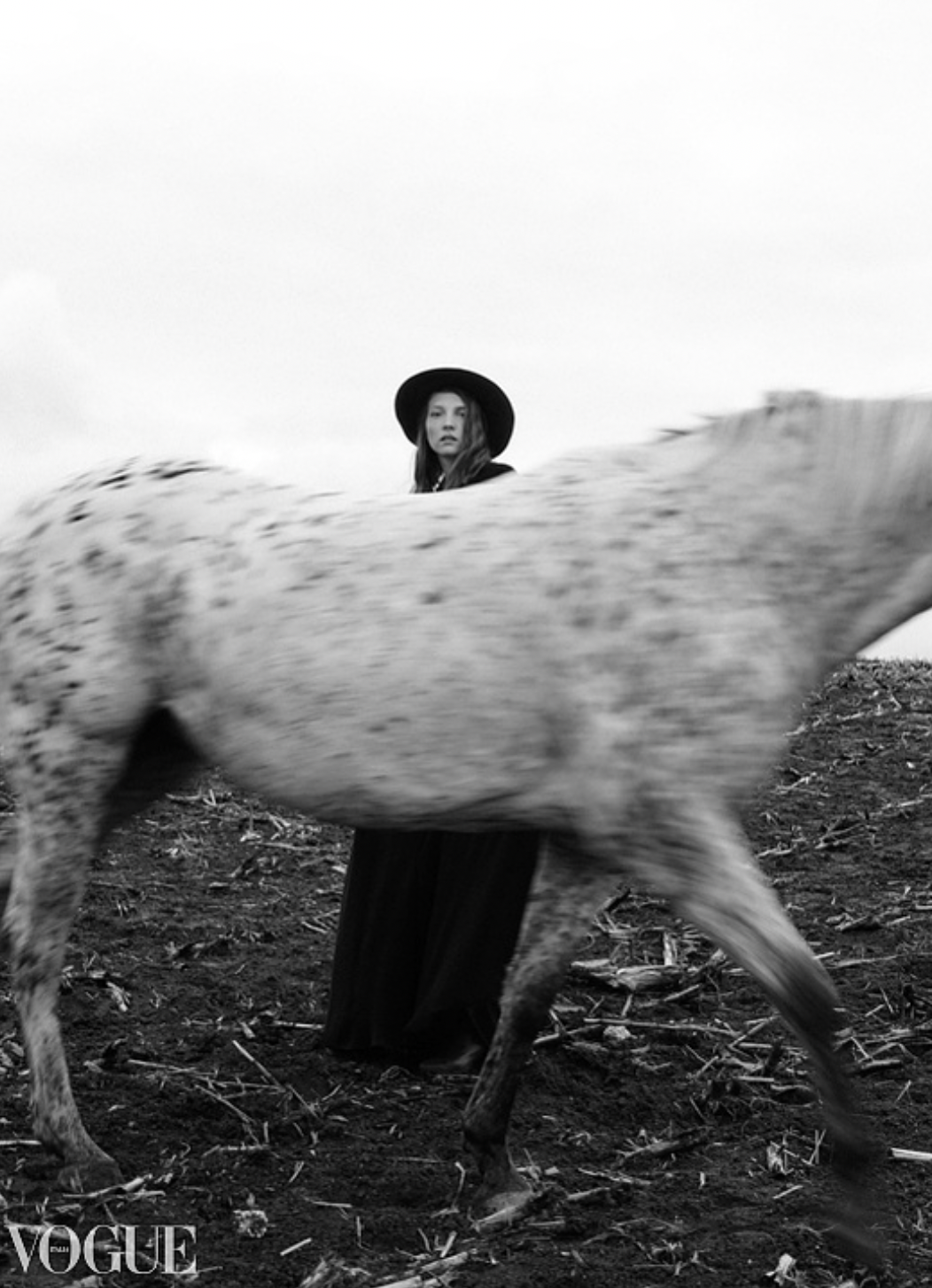 Black and white Vogue editorial of woman in black hat standing behind a blurred spotted horse.