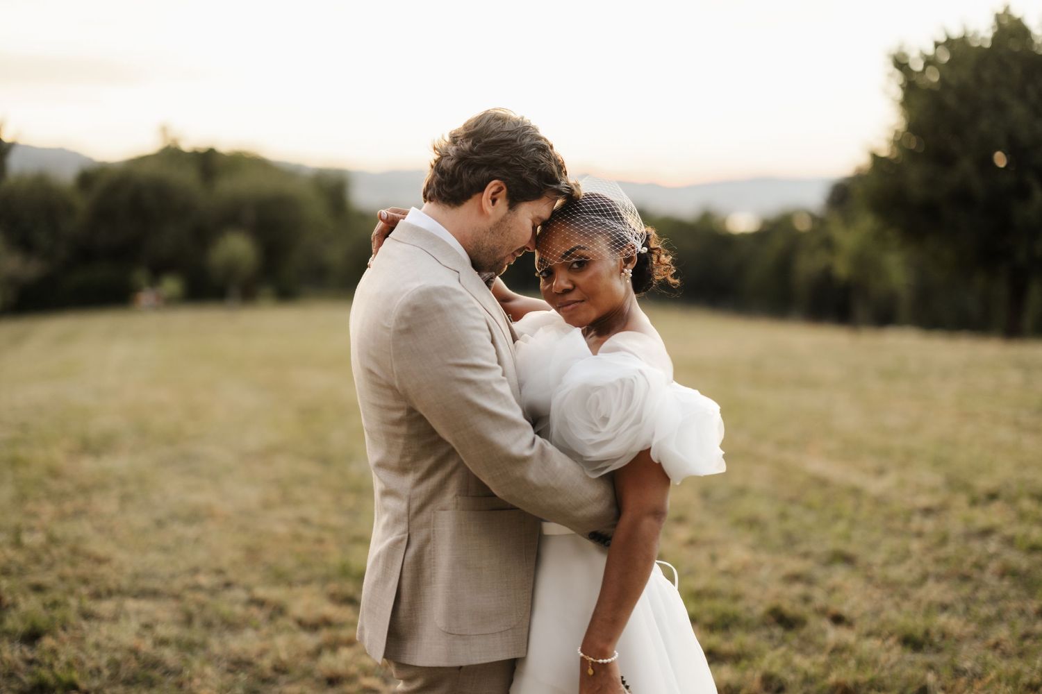 Interracial wedding couple sharing a tender embrace in a sunlit meadow, bride in white dress, groom in beige suit.