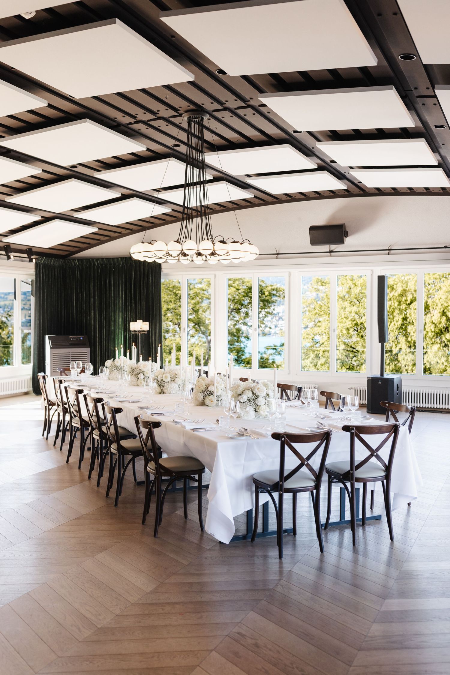 Elegant wedding reception hall with long white-draped tables, wooden cross-back chairs, herringbone floors, and a modern chandelier.