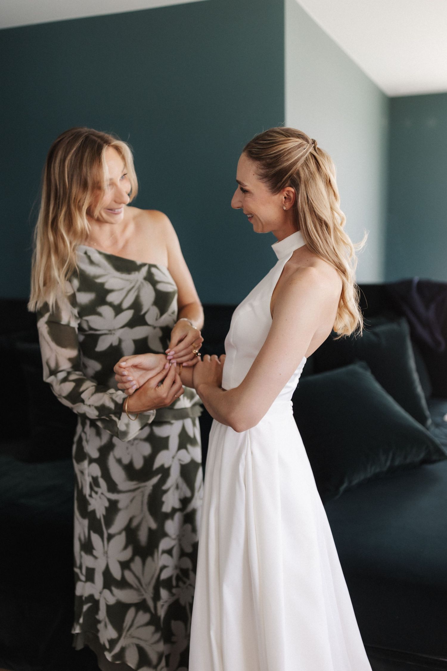 Two blonde women smile at each other, one in a white wedding gown and one in a floral dress, holding hands indoors.
