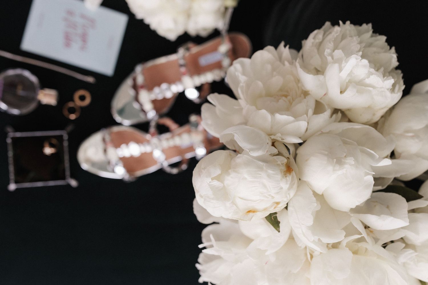 White peony bouquet with rose gold heels, perfume, and wedding accessories on black.
