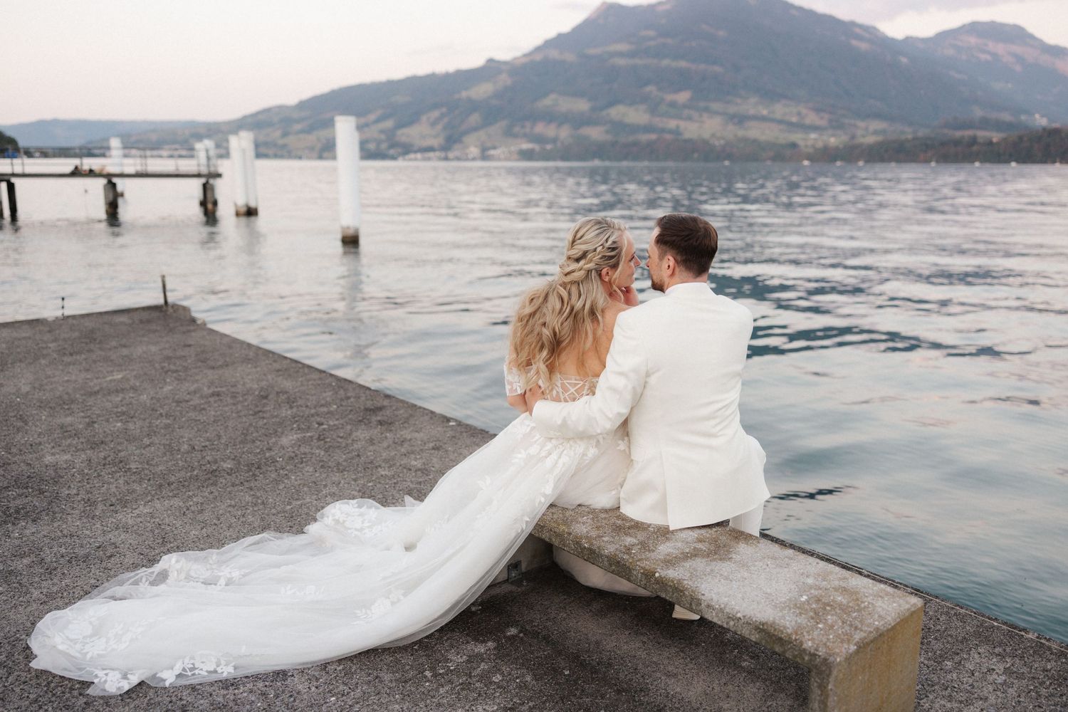 Wedding couple sitting on a lakeside dock with sweeping mountain scenery and long bridal train displayed.