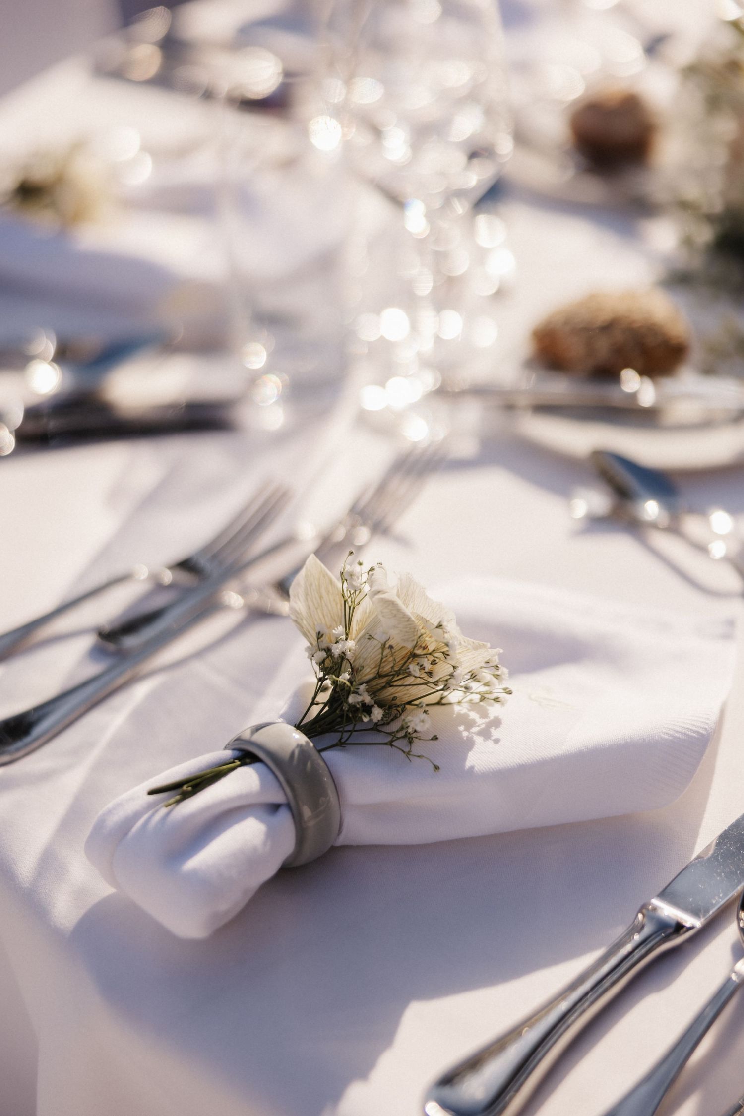 A white folded napkin adorned with white roses and baby's breath sits on a elegant wedding table setting.