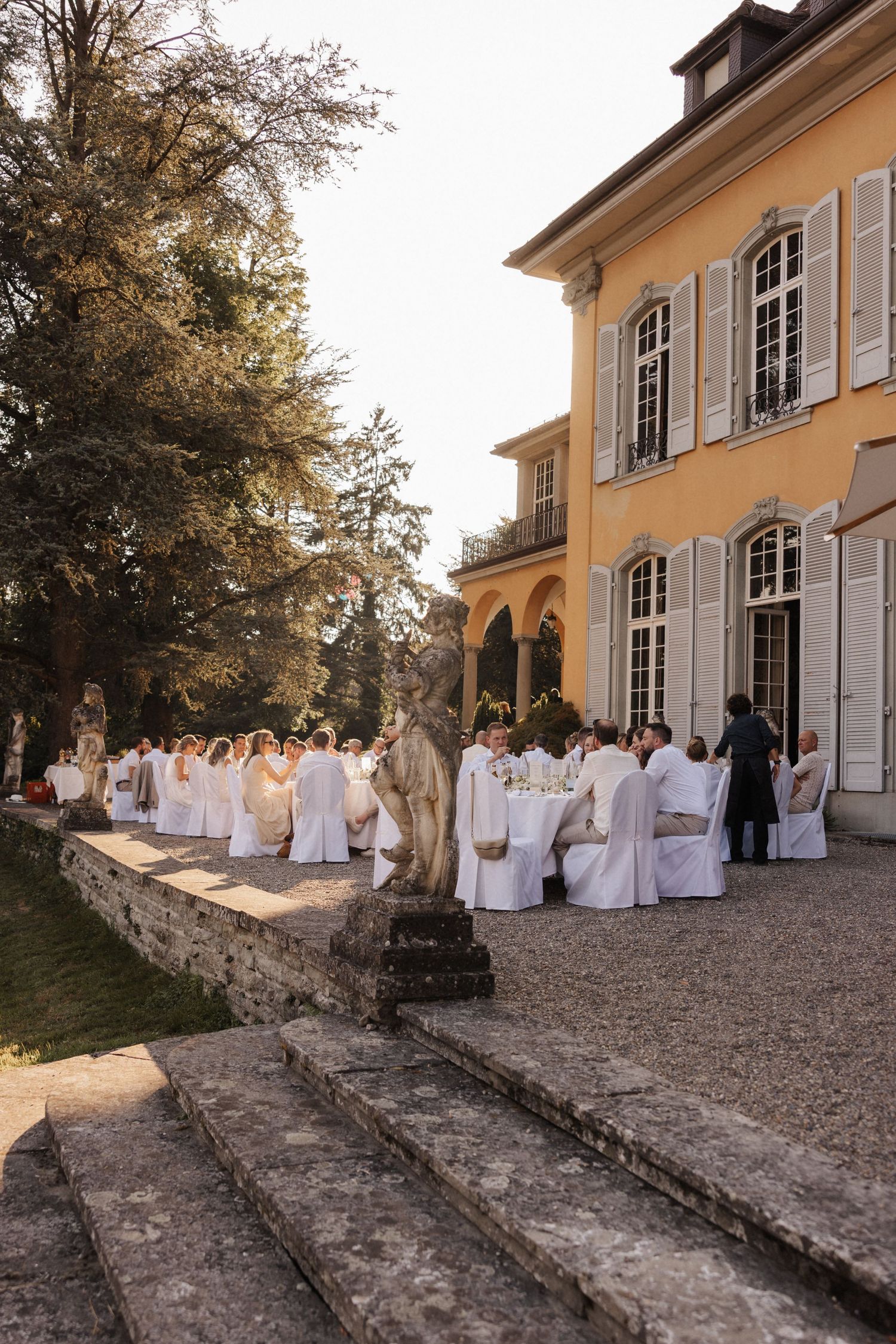 Outdoor wedding reception beside a yellow European manor with stone statues and guests dressed in white at dusk.