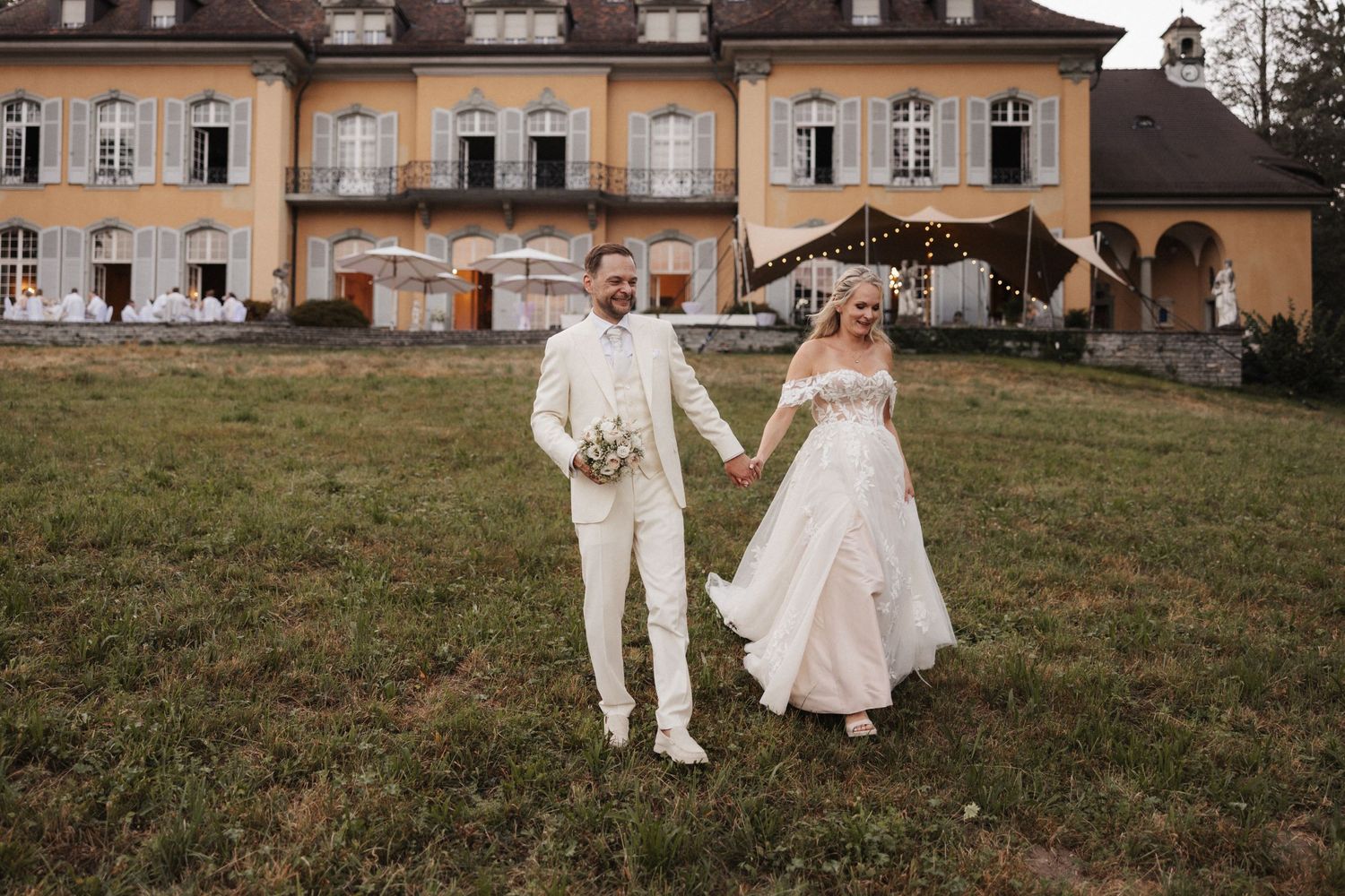Bride and groom holding hands walking across a green lawn in front of a yellow European manor house.