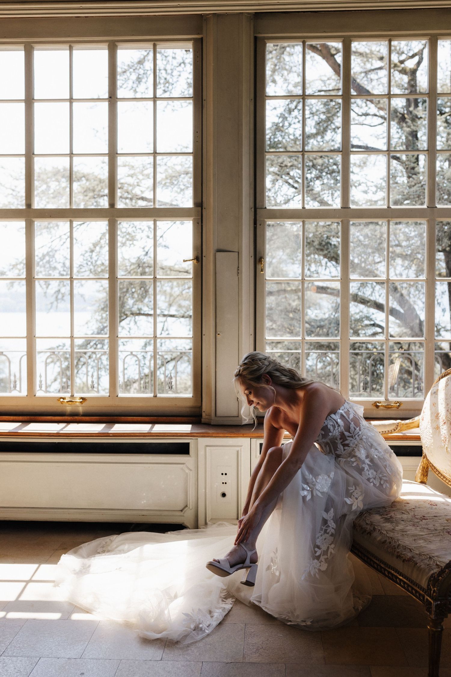Bride in floral lace wedding gown putting on heels while seated by large sunlit windows.