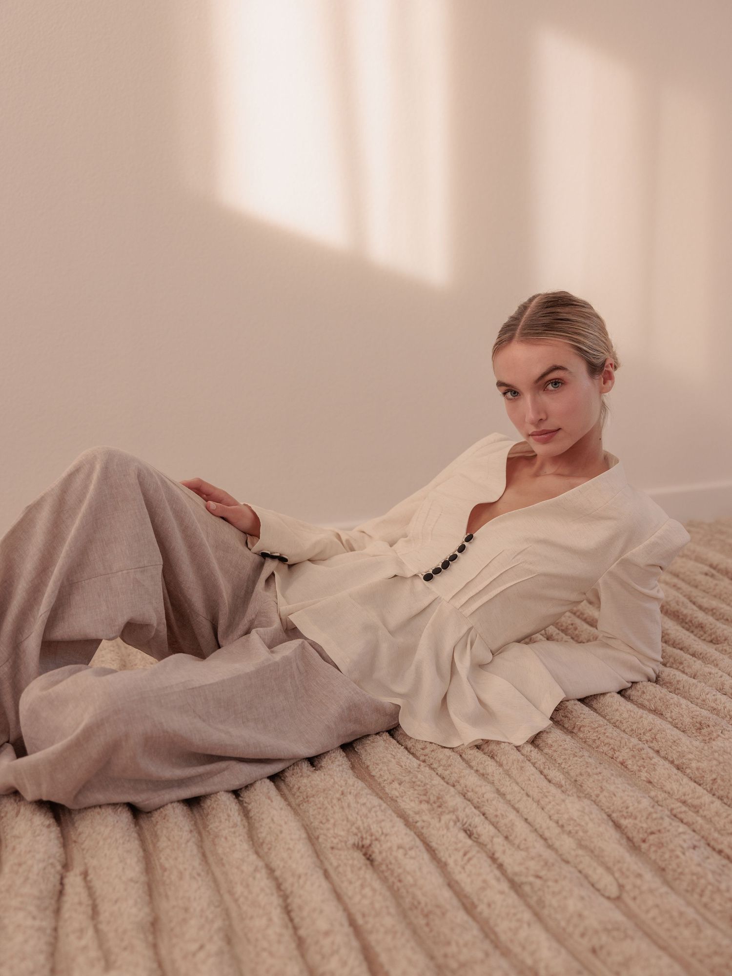 Woman in cream blouse and grey trousers reclines gracefully on a textured surface in soft natural light.