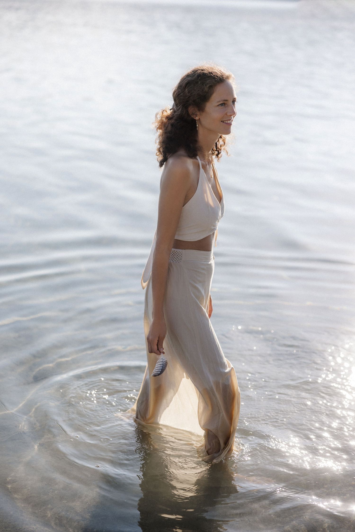 Smiling woman in white crop top and beige skirt walking gracefully through shallow water.