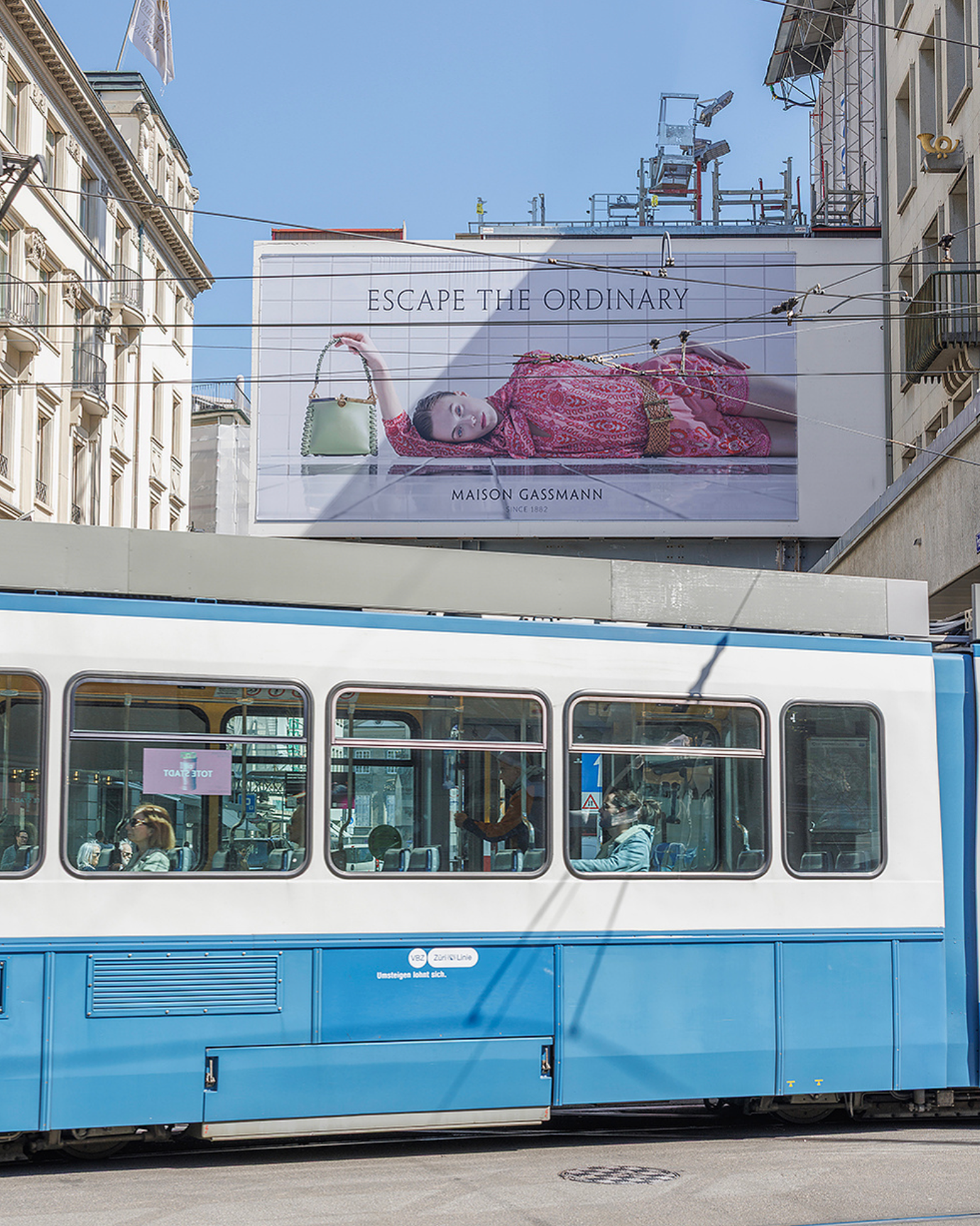 Blue city tram passing billboard with Escape the Ordinary fashion advertisement on urban street.
