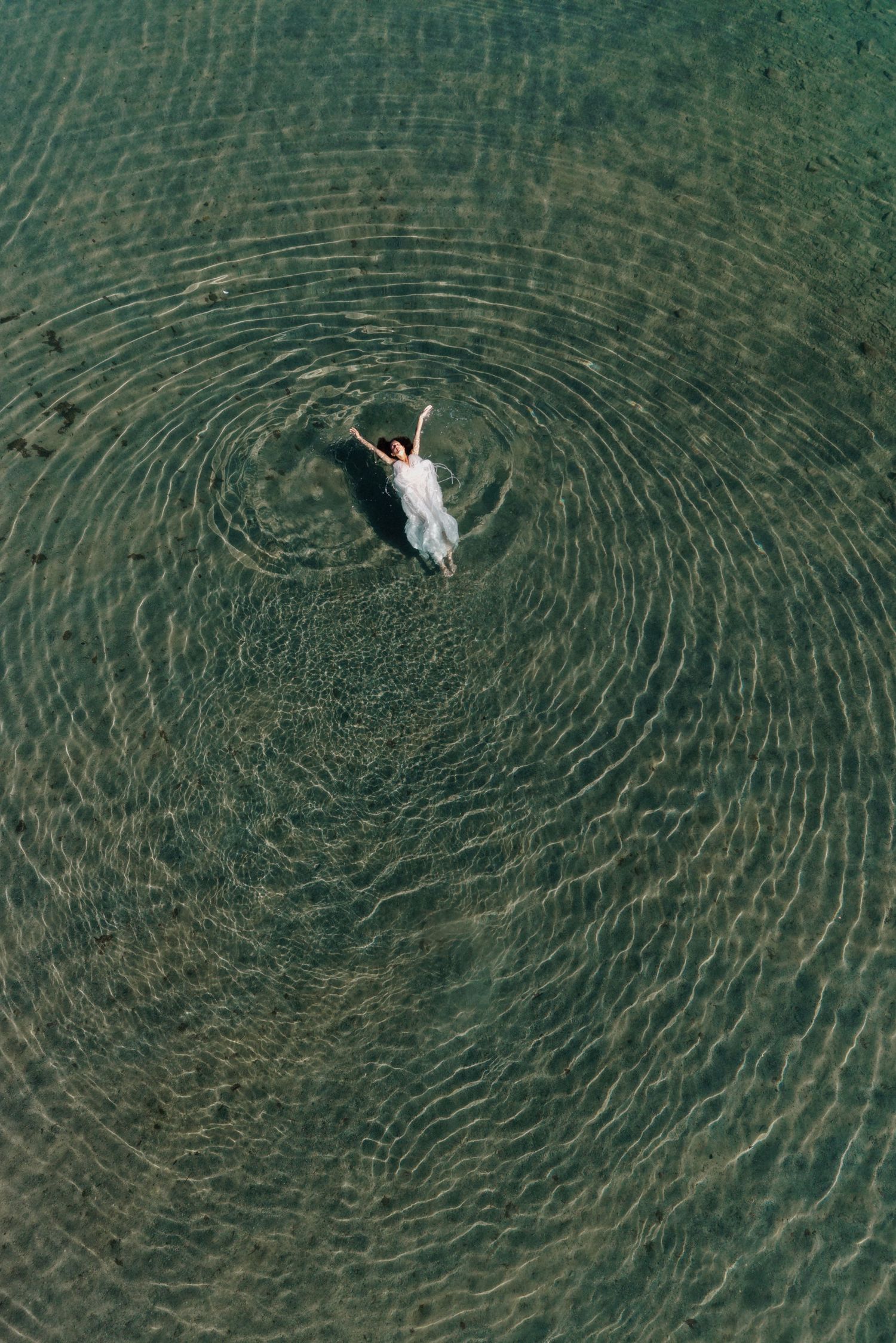 Aerial view of a woman in a white dress floating in clear turquoise water creating circular ripples.