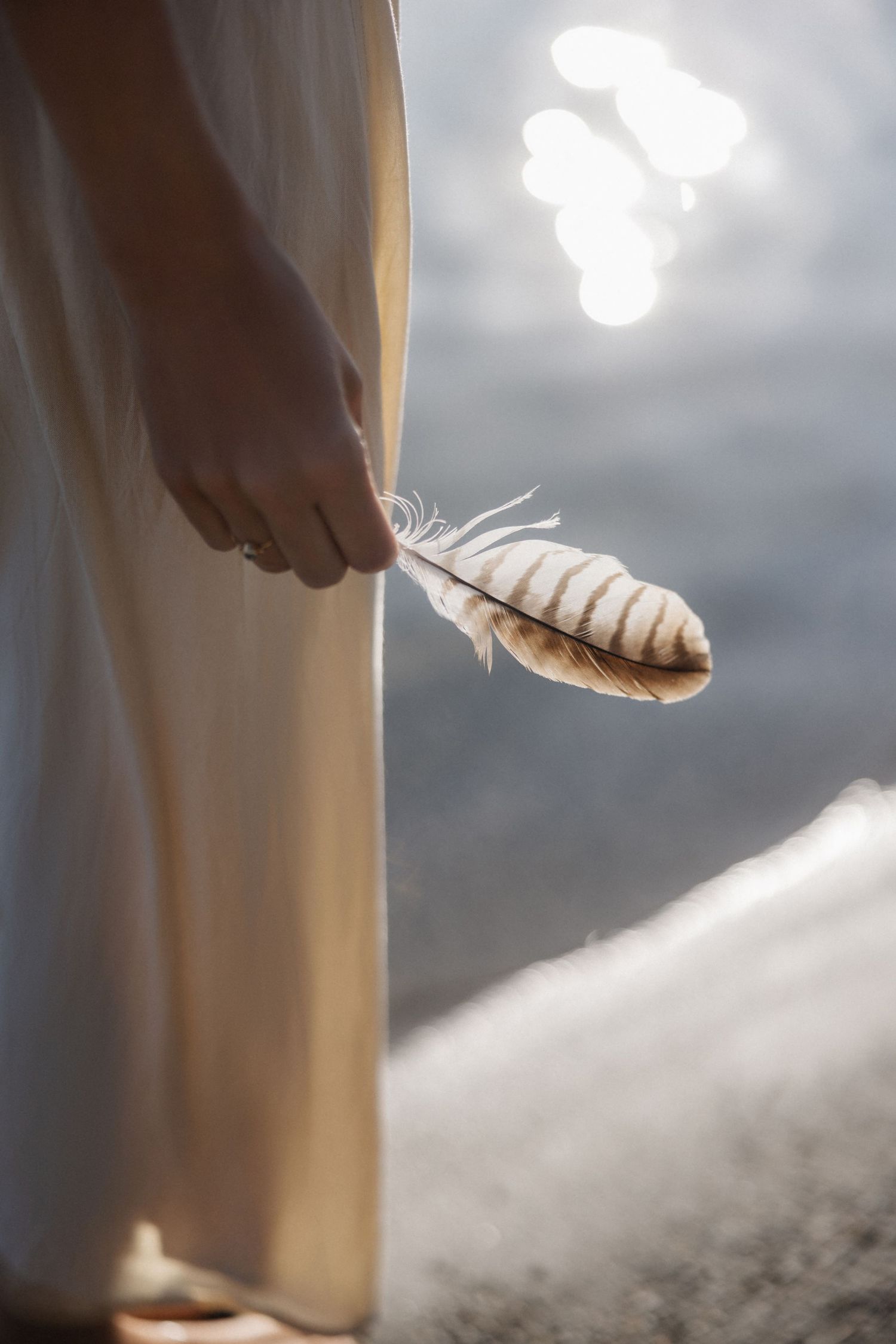 Close-up of woman's hand holding a striped feather beside a flowing cream dress near sparkling water.