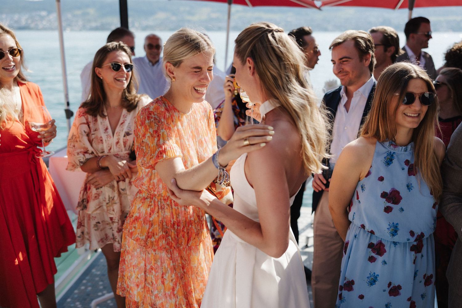 Elegantly dressed wedding guests mingling and dancing at a sunny outdoor lakeside reception.