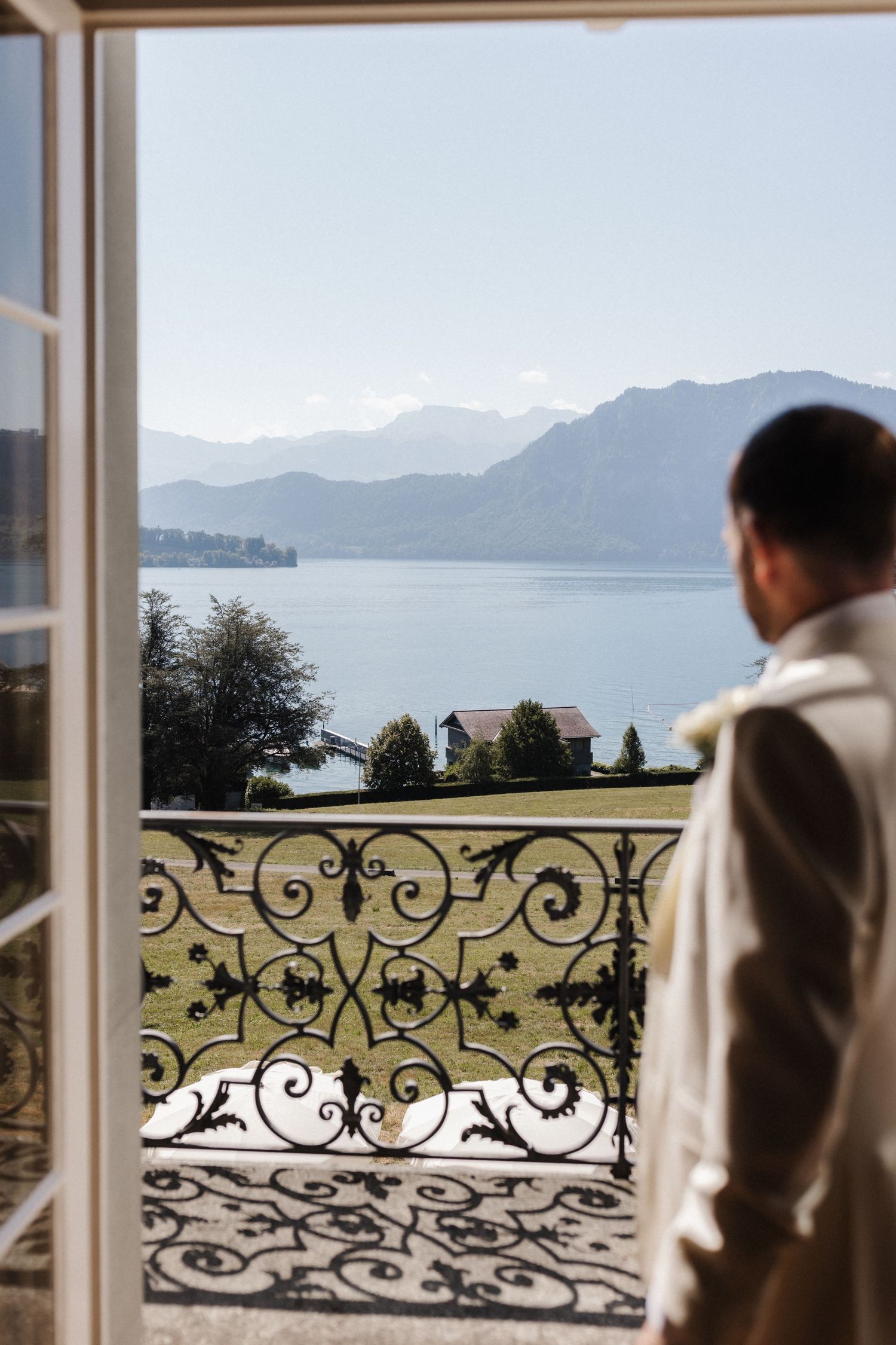 Groom in white suit gazes at stunning lake and mountain view through ornate balcony doors.