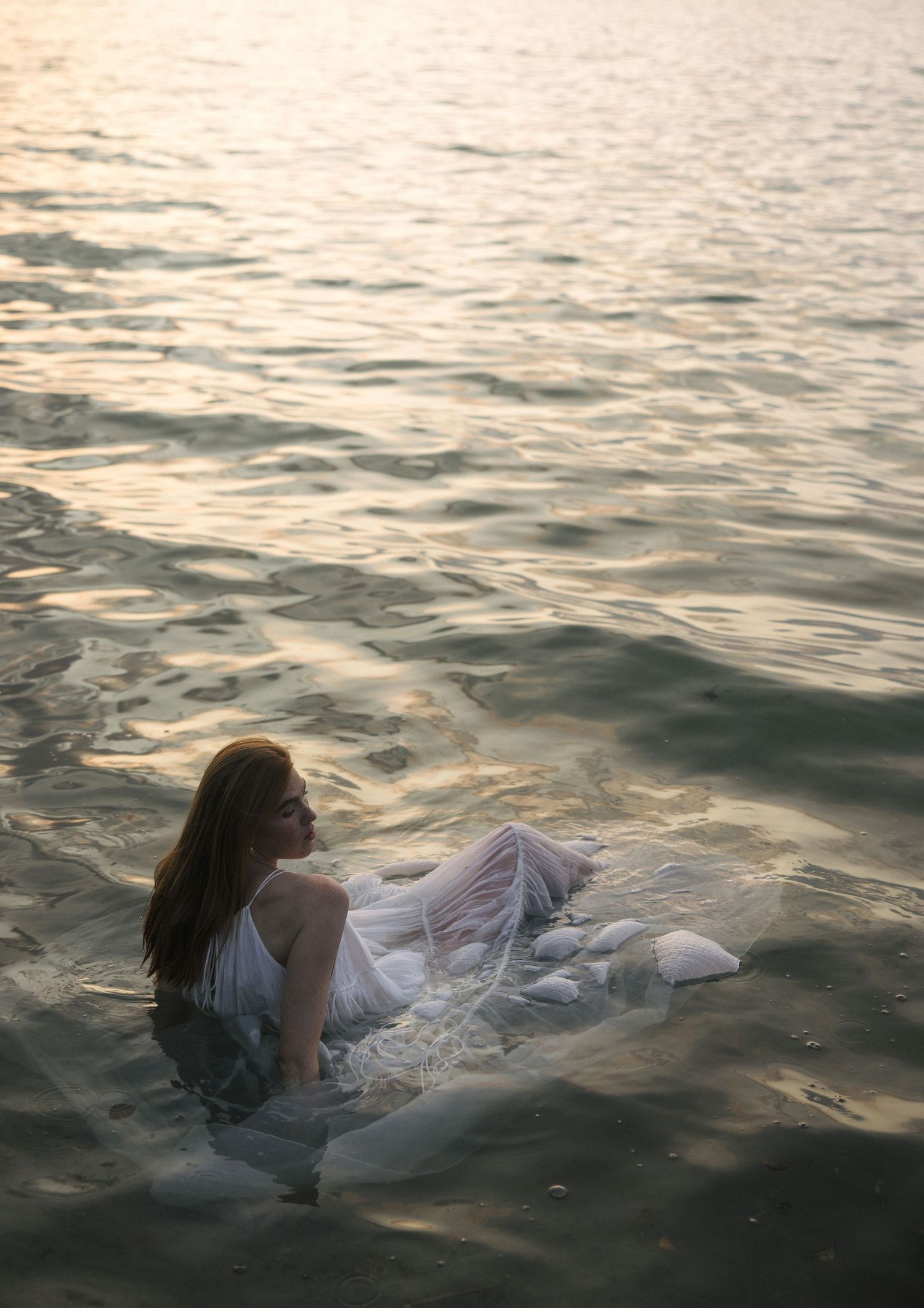 Woman in white dress sitting in shallow golden water at sunset by the shore.