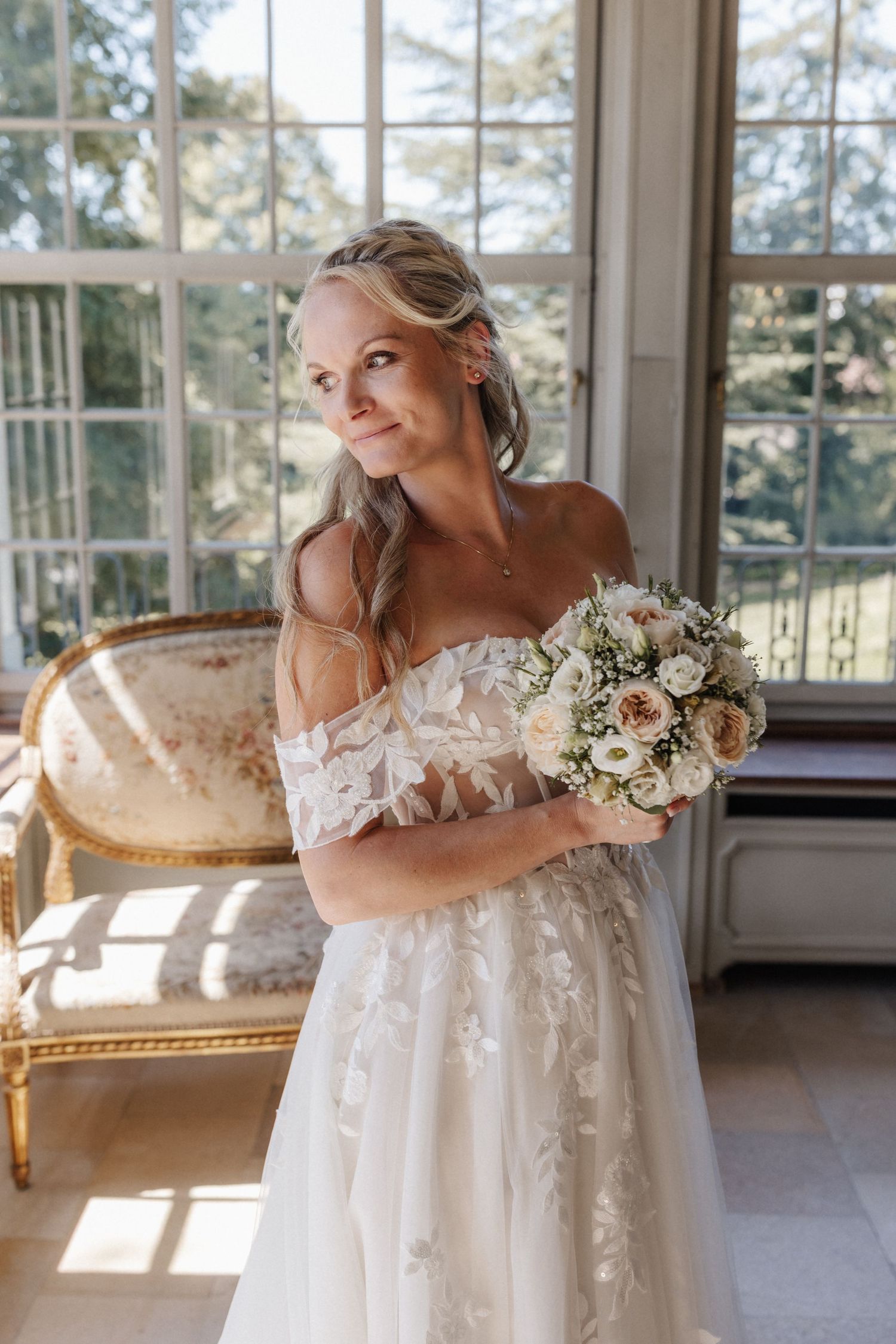 Smiling bride in off-shoulder lace gown holding white and blush bouquet in a sunlit elegant room.
