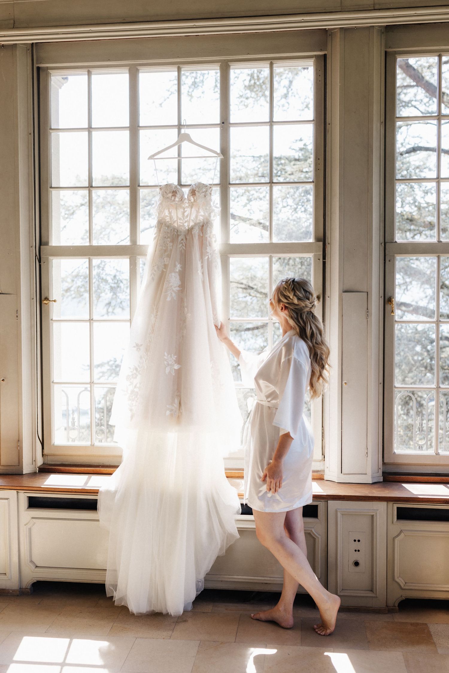 A bride in a white robe reaching for her lace wedding dress hanging by a large window.