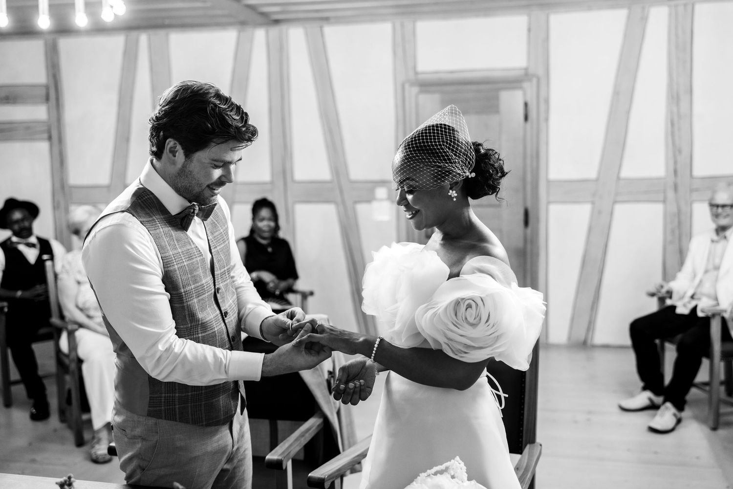 Groom placing wedding ring on brides finger during interracial wedding ceremony in black and white.