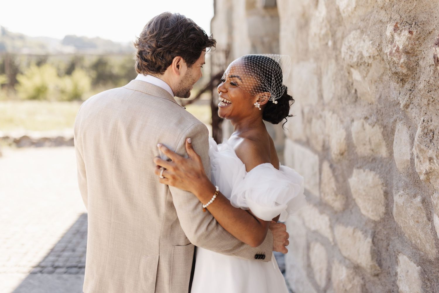 Happy interracial bride and groom embracing and smiling against rustic stone wall after wedding.