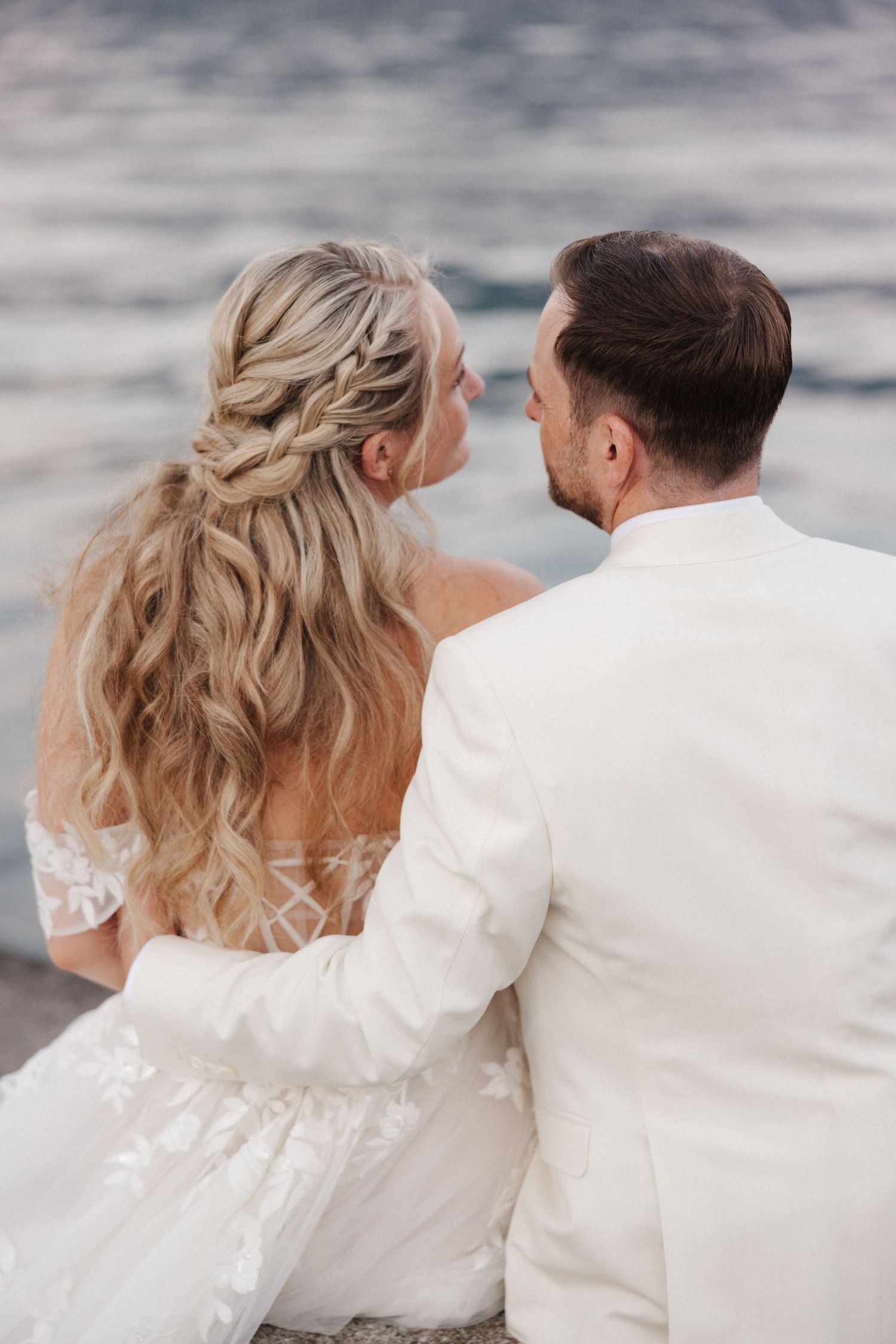 Rear view of bride and groom sitting by the water, bride showing elegant braided hairstyle and lace dress.