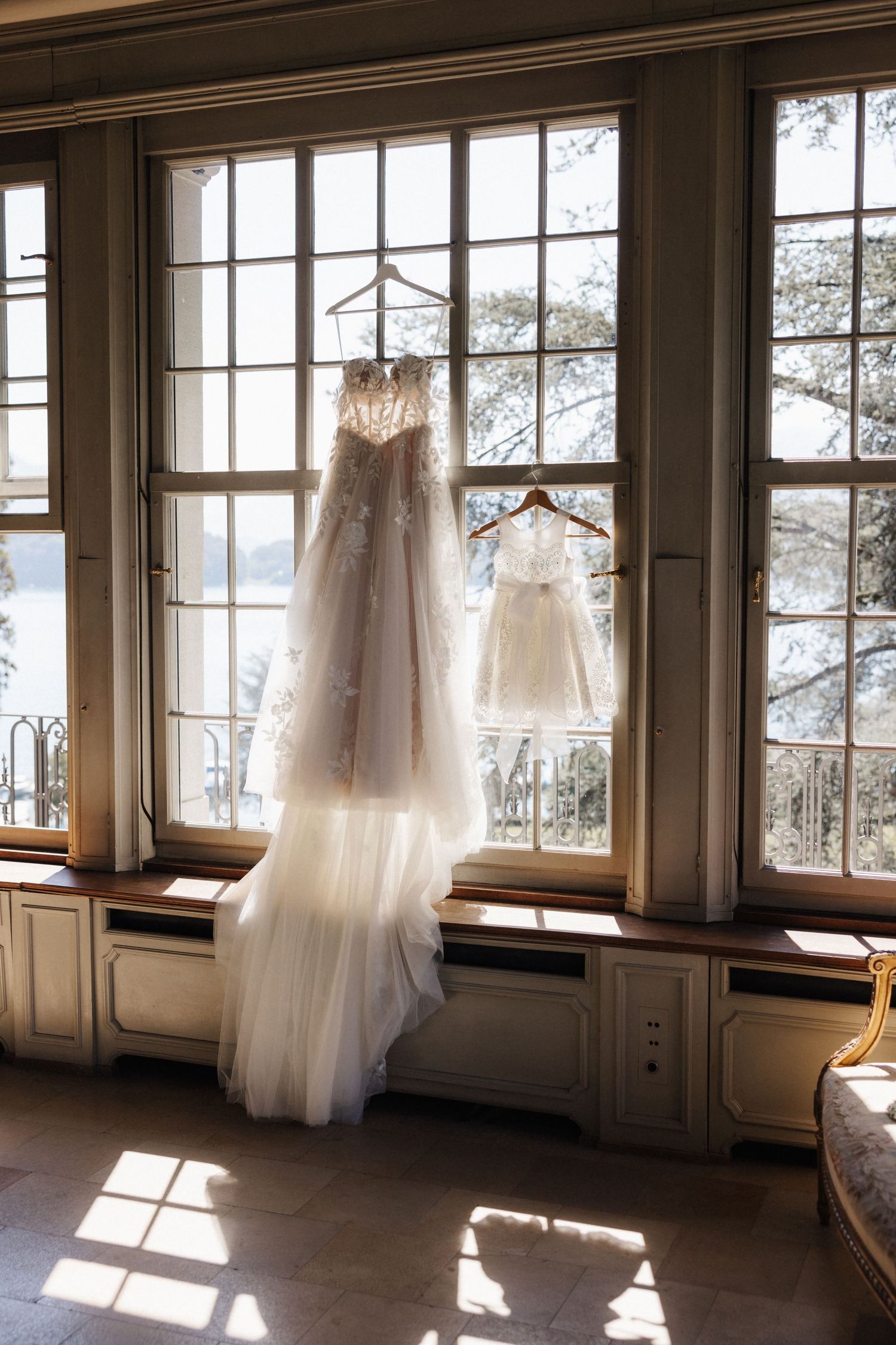 A lace wedding gown and flower girl dress hanging on a window with a scenic view outside.