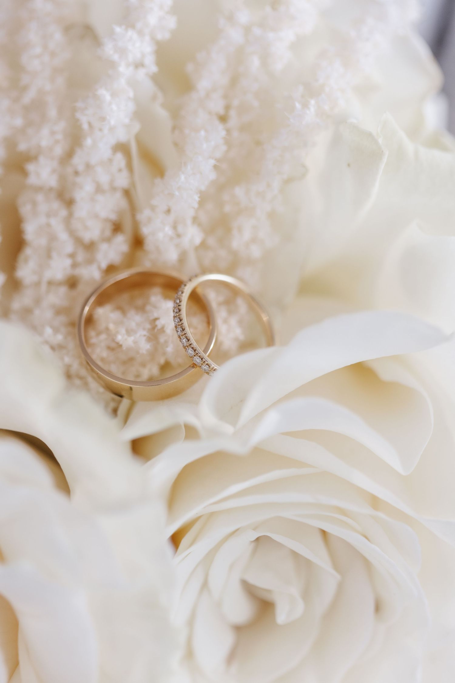 Two gold wedding bands, one with diamonds, placed on white roses and soft white floral details.