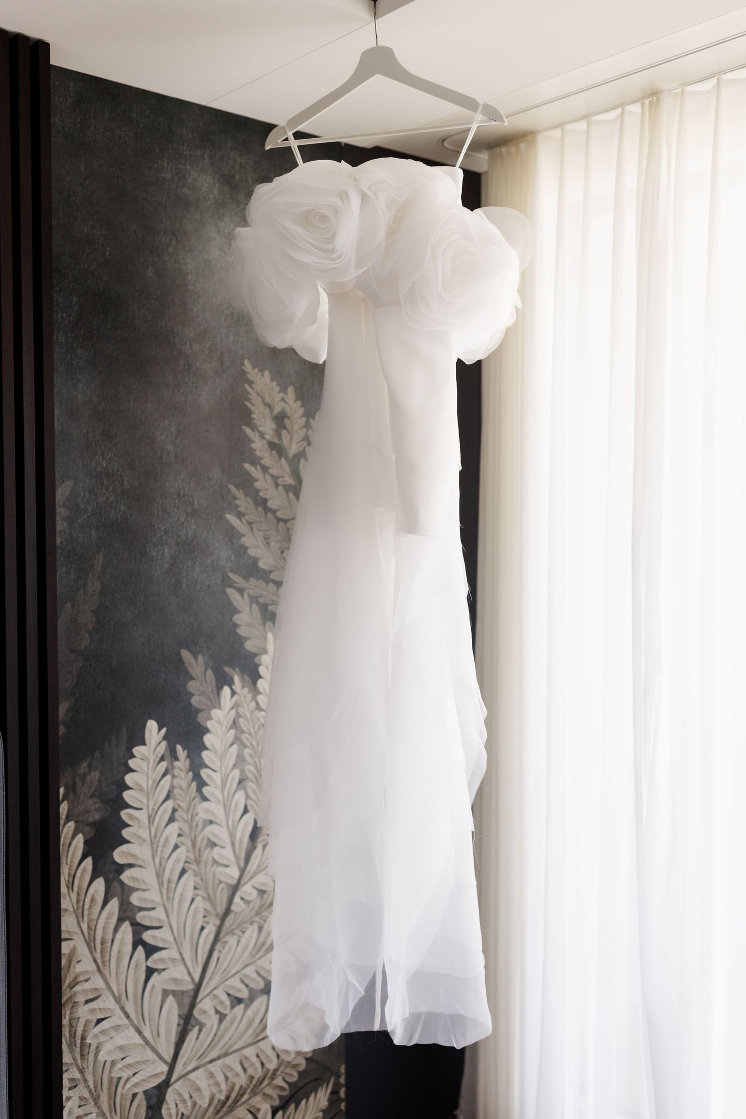 Elegant white strapless wedding dress with oversized organza roses hanging against decorative wall.