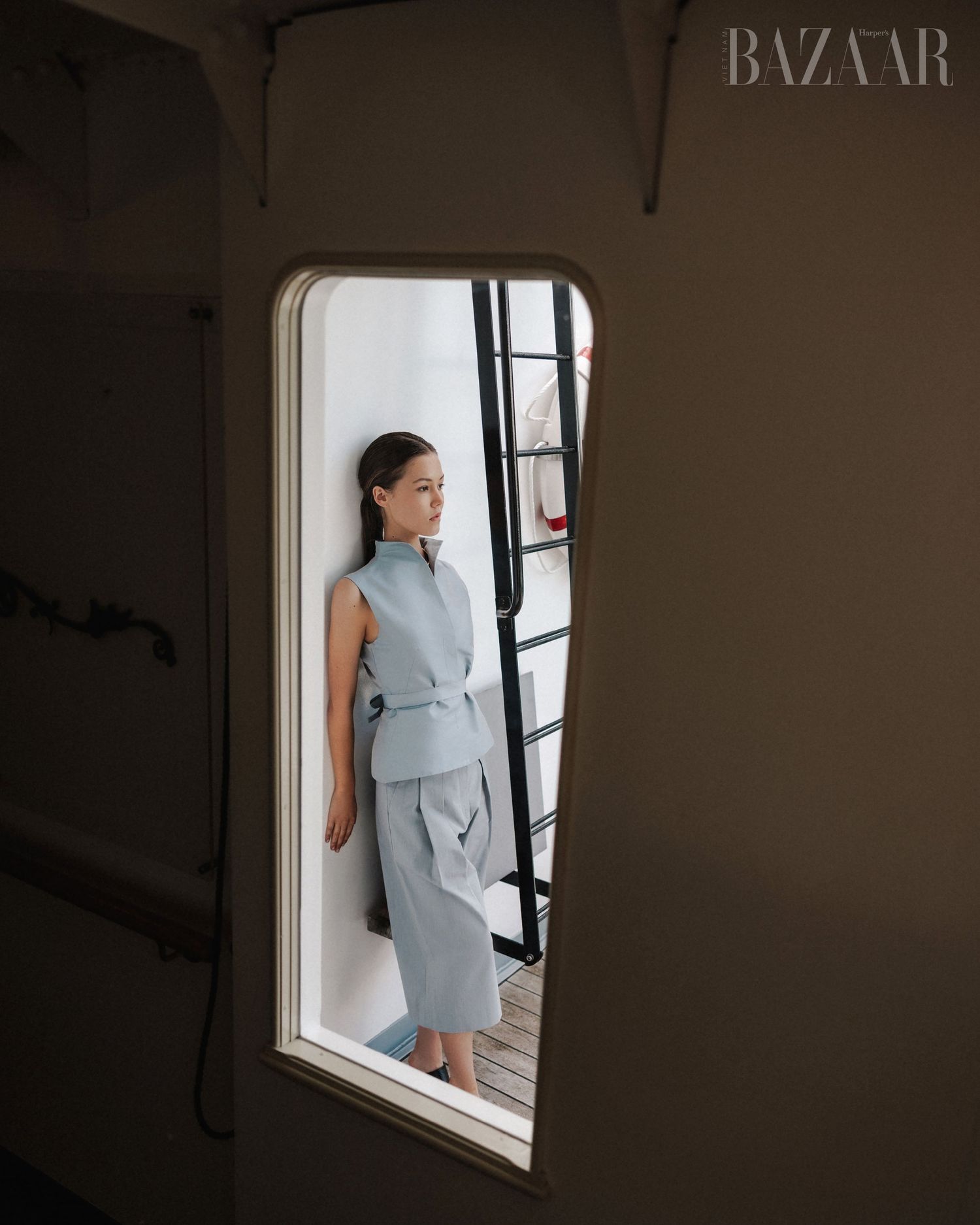 Model framed in oval window wearing powder blue sleeveless suit, minimalist fashion editorial for Harper's Bazaar.