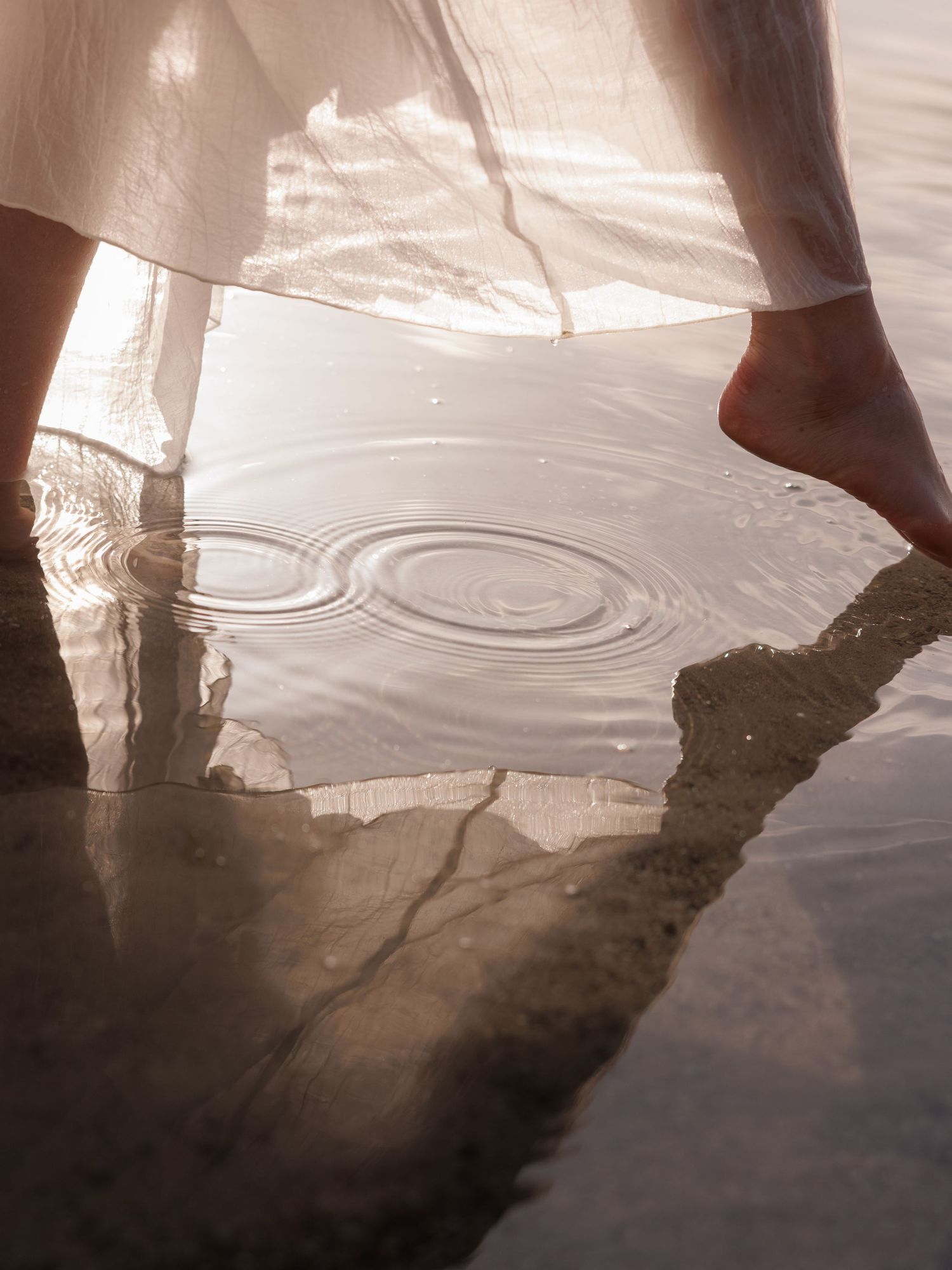 Barefoot woman in white dress standing in shallow water creating circular ripples.