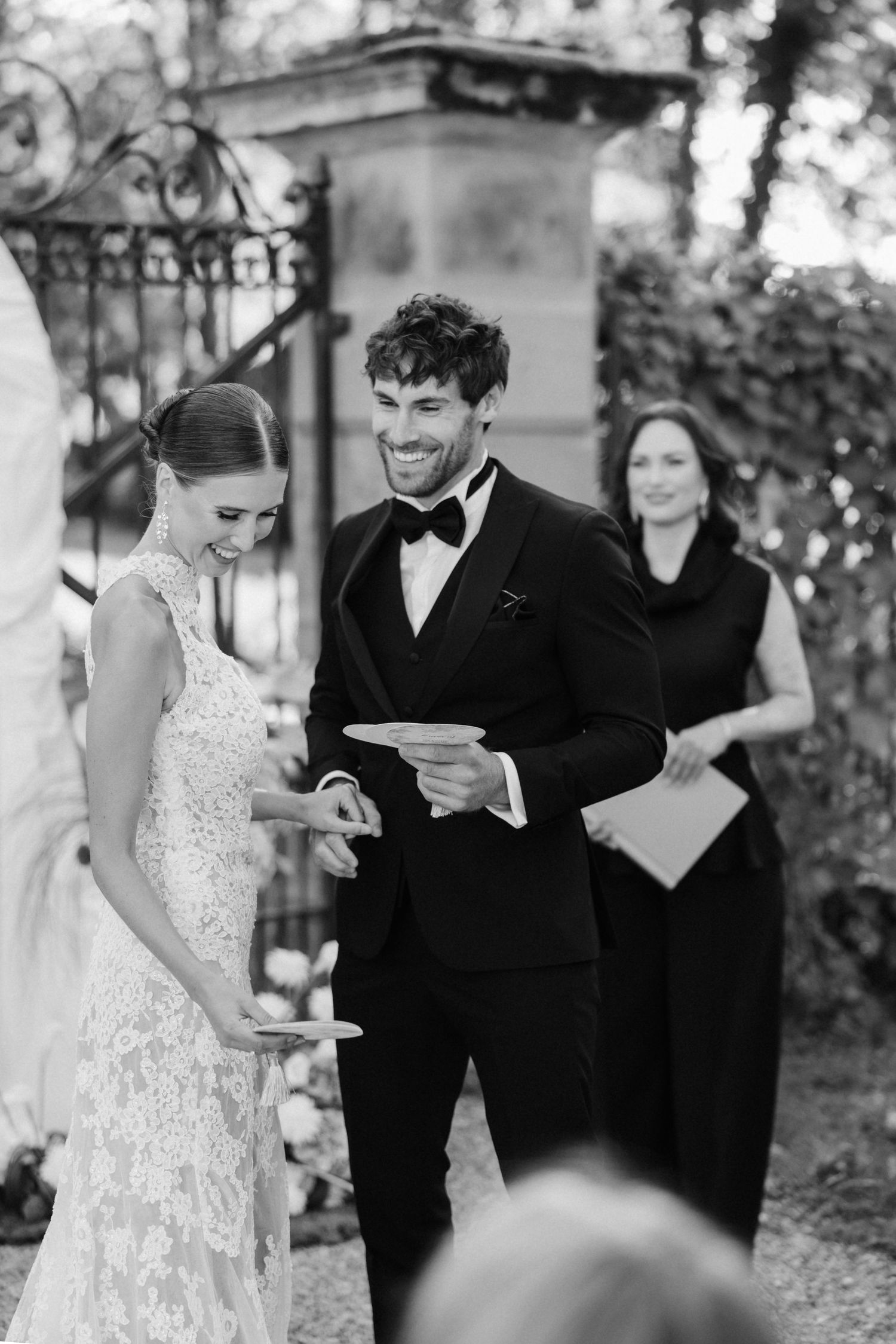 Black and white photo of smiling bride and groom exchanging rings at outdoor wedding ceremony.